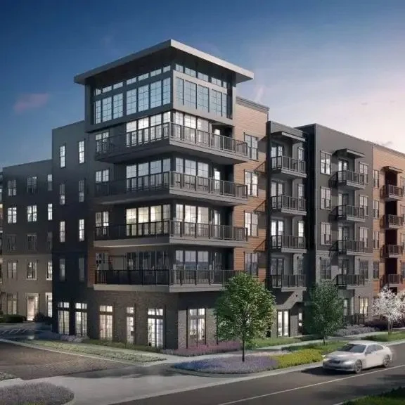 Modern multi-story apartment building with large glass windows and balconies, surrounded by a sidewalk, trees, and a street with cars, under a blue sky.