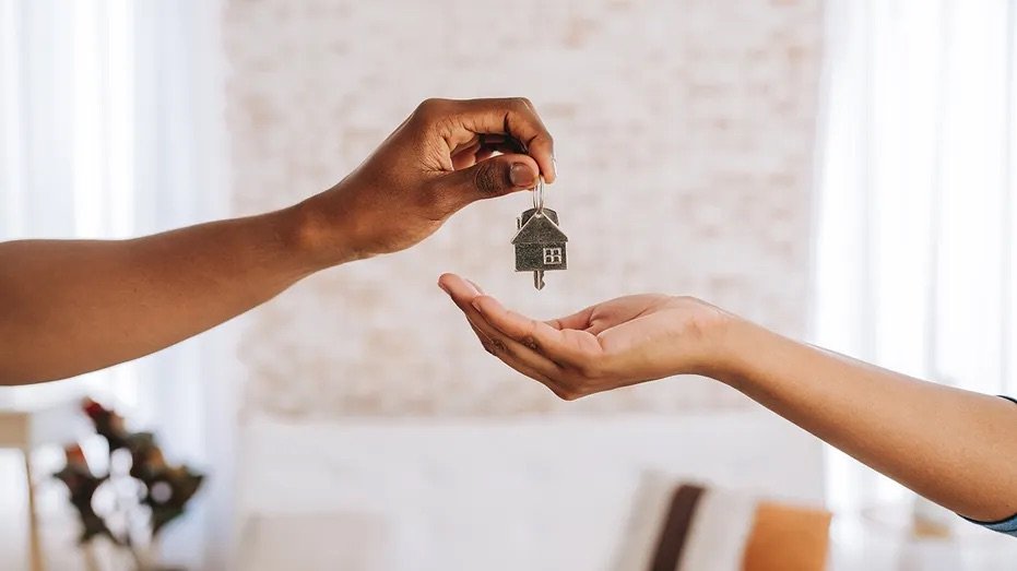 One person is handing a key with a house-shaped keychain to another person inside a home.
