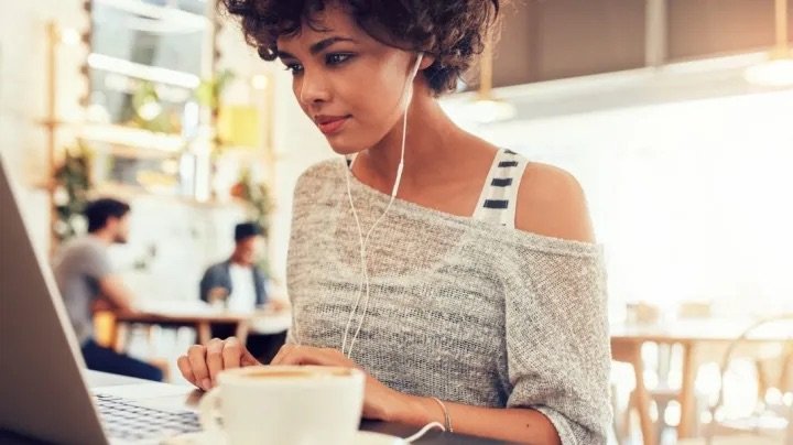 A young woman with curly hair using a laptop in a cafe, wearing earphones and a casual top.