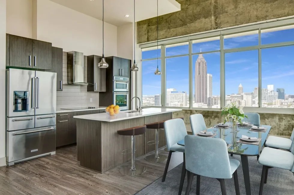 Modern kitchen and dining area with large windows overlooking a city skyline, including the Empire State Building, with stainless steel appliances, a kitchen island with stools, and a table set for six with a flower centerpiece.