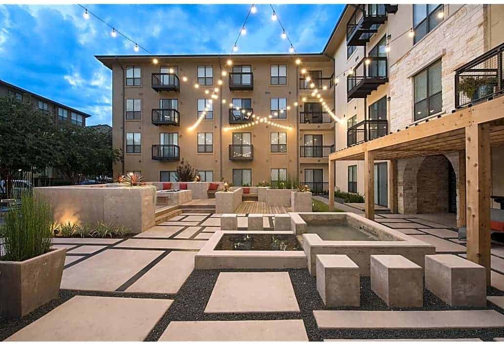 An outdoor courtyard of an apartment complex at dusk, decorated with string lights, featuring seating areas, plants, a small pool or fountain, and modern architectural elements.
