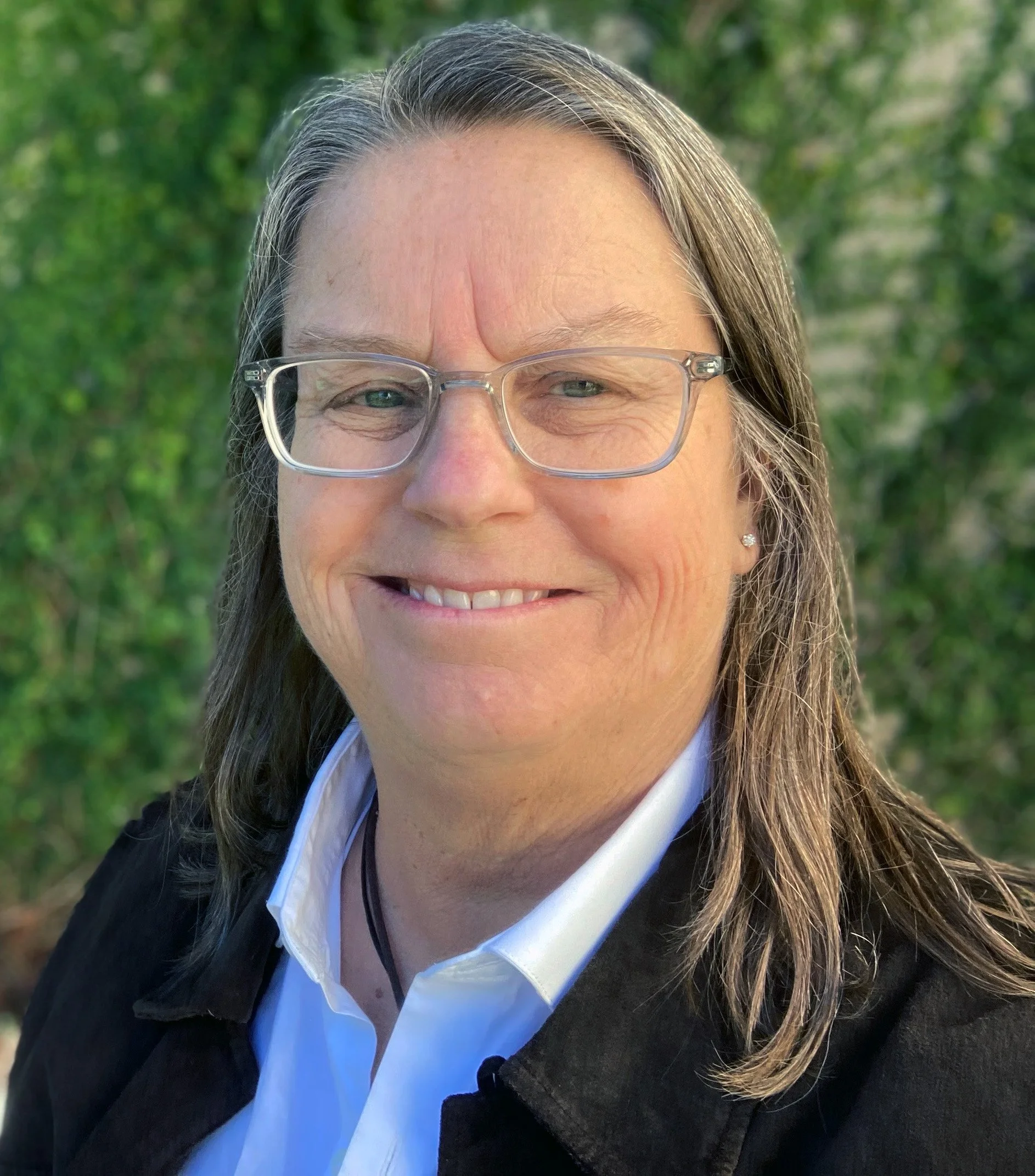 Picture of Maja Gray with long brown hair and glasses smiling outdoors with green trees in the background.