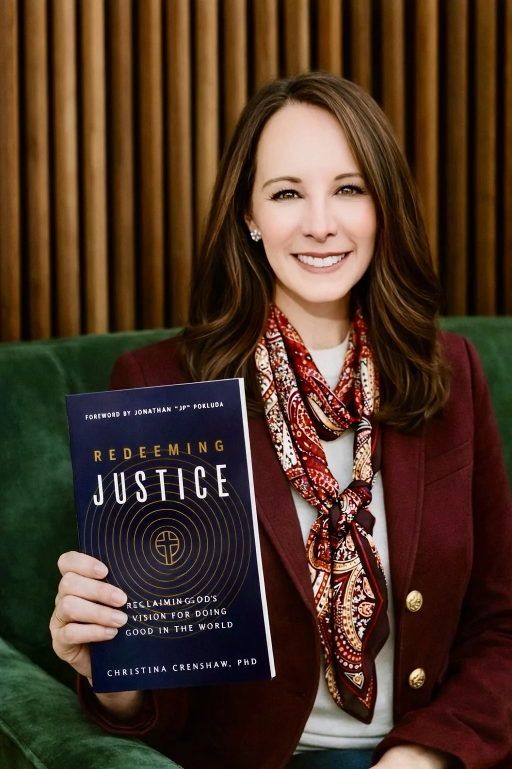 A woman with long brown hair smiling, wearing a red blazer and a patterned scarf, sitting on a green couch in front of wooden paneling, holding a book titled "Redeeming Justice" by Christina Crenshaw, PhD.