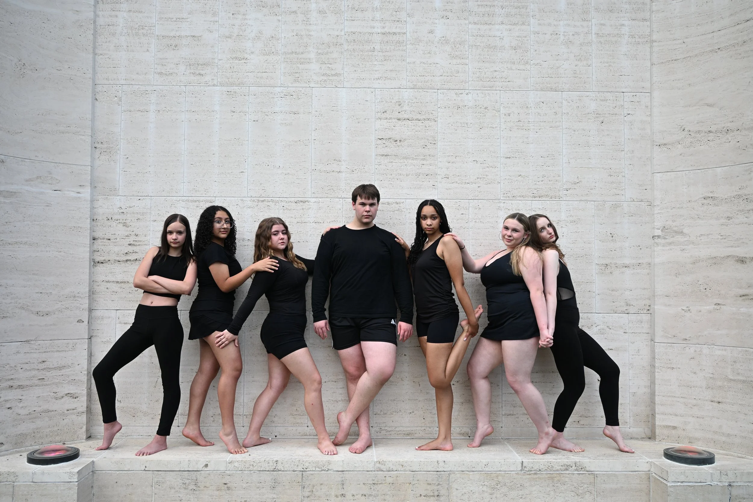 A group of seven diverse young women standing in front of a large light-colored stone wall, with some barefoot, posing confidently.