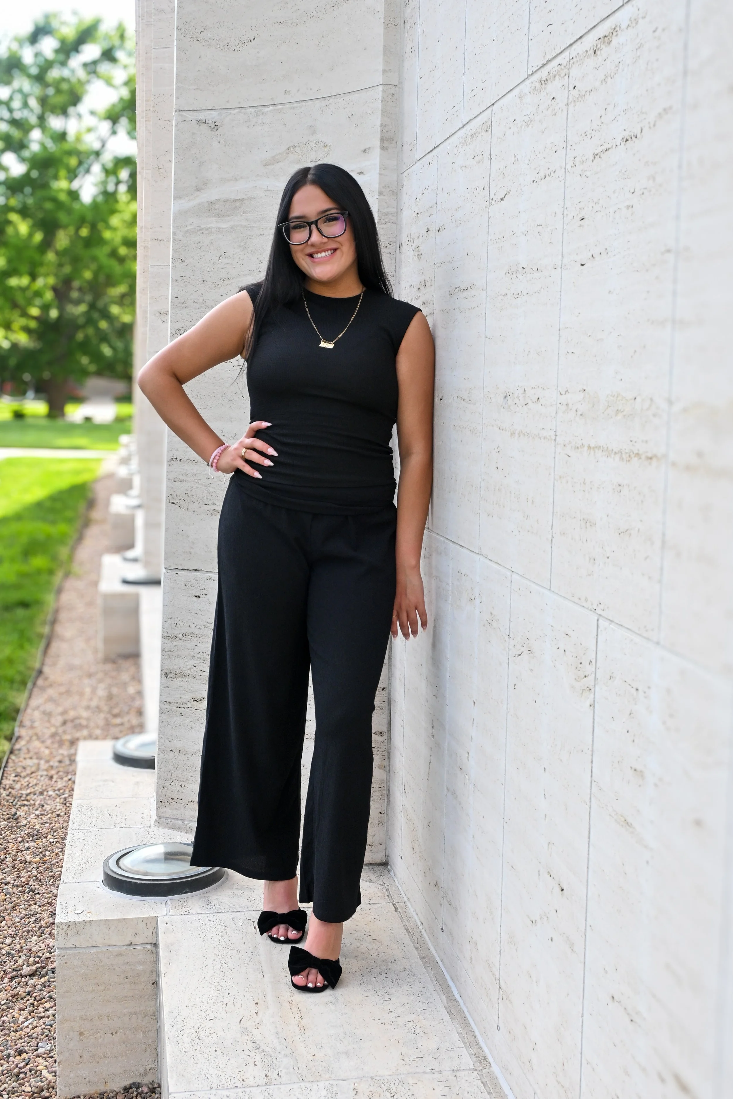 A woman with long black hair, glasses, and a big smile standing outdoors against a light-colored stone wall, dressed in black sleeveless top and pants, wearing black high-heeled shoes with bows, and accessories including a necklace and bracelet, with green trees blurred in the background.