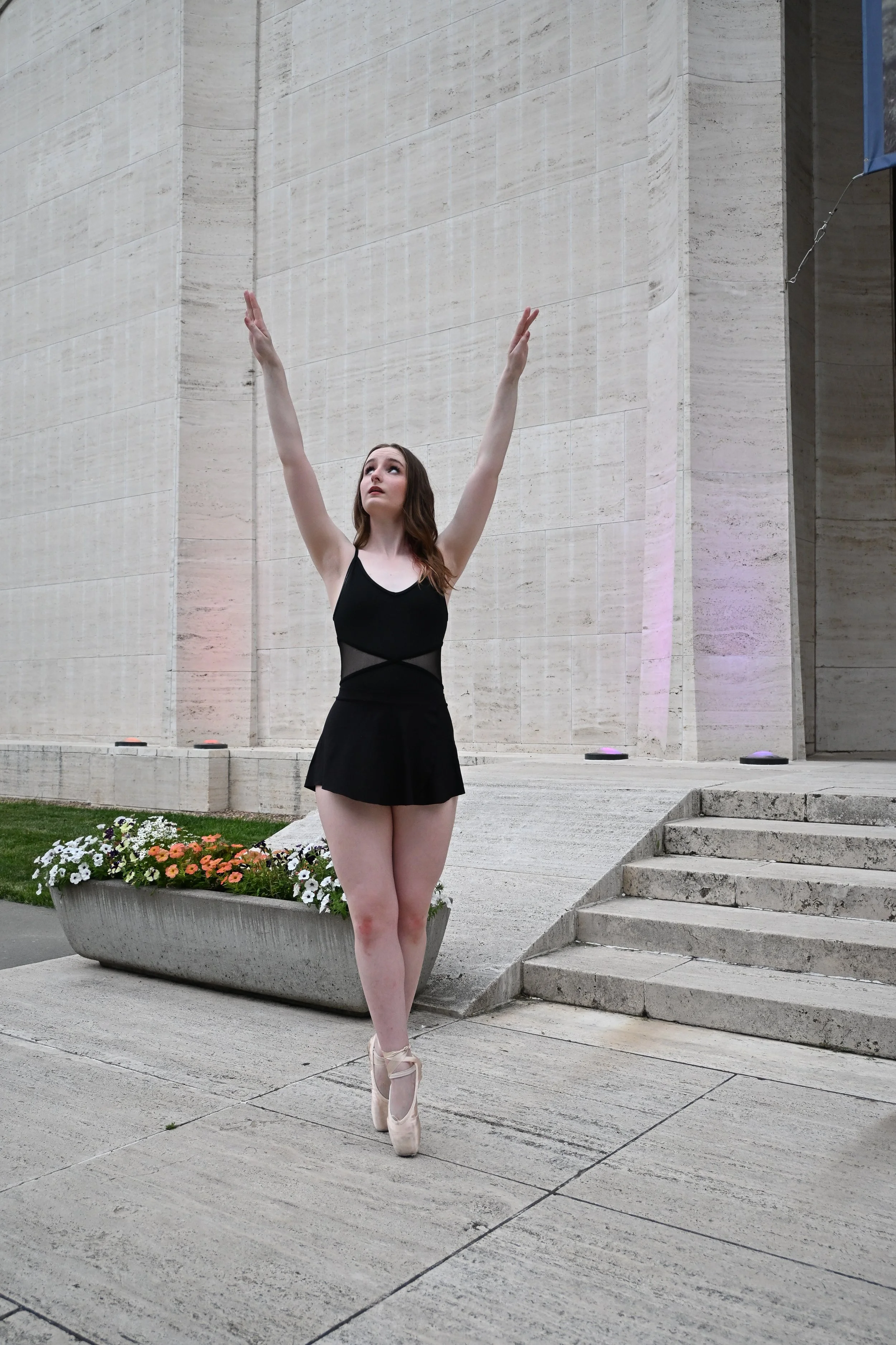 A ballet dancer in a black leotard and pink tutu standing on pointe outside of a large stone building with stairs and flower planter.