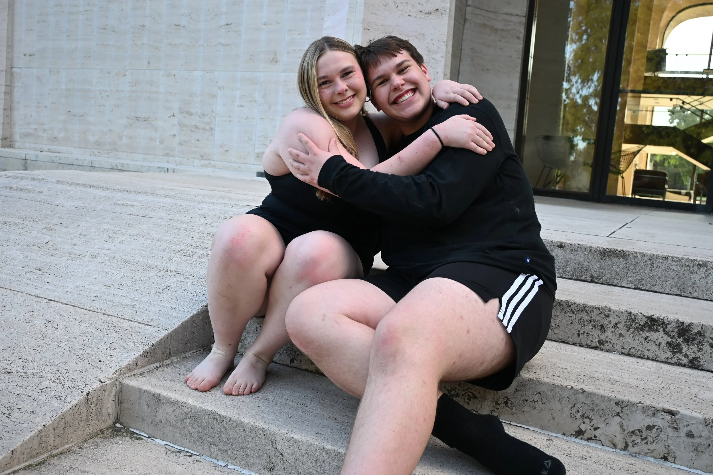 Two young people, a girl and a boy, sitting on steps outside, hugging and smiling at the camera. The girl has long blonde hair and is wearing a black dress, while the boy has short dark hair and is wearing a black shirt and shorts.