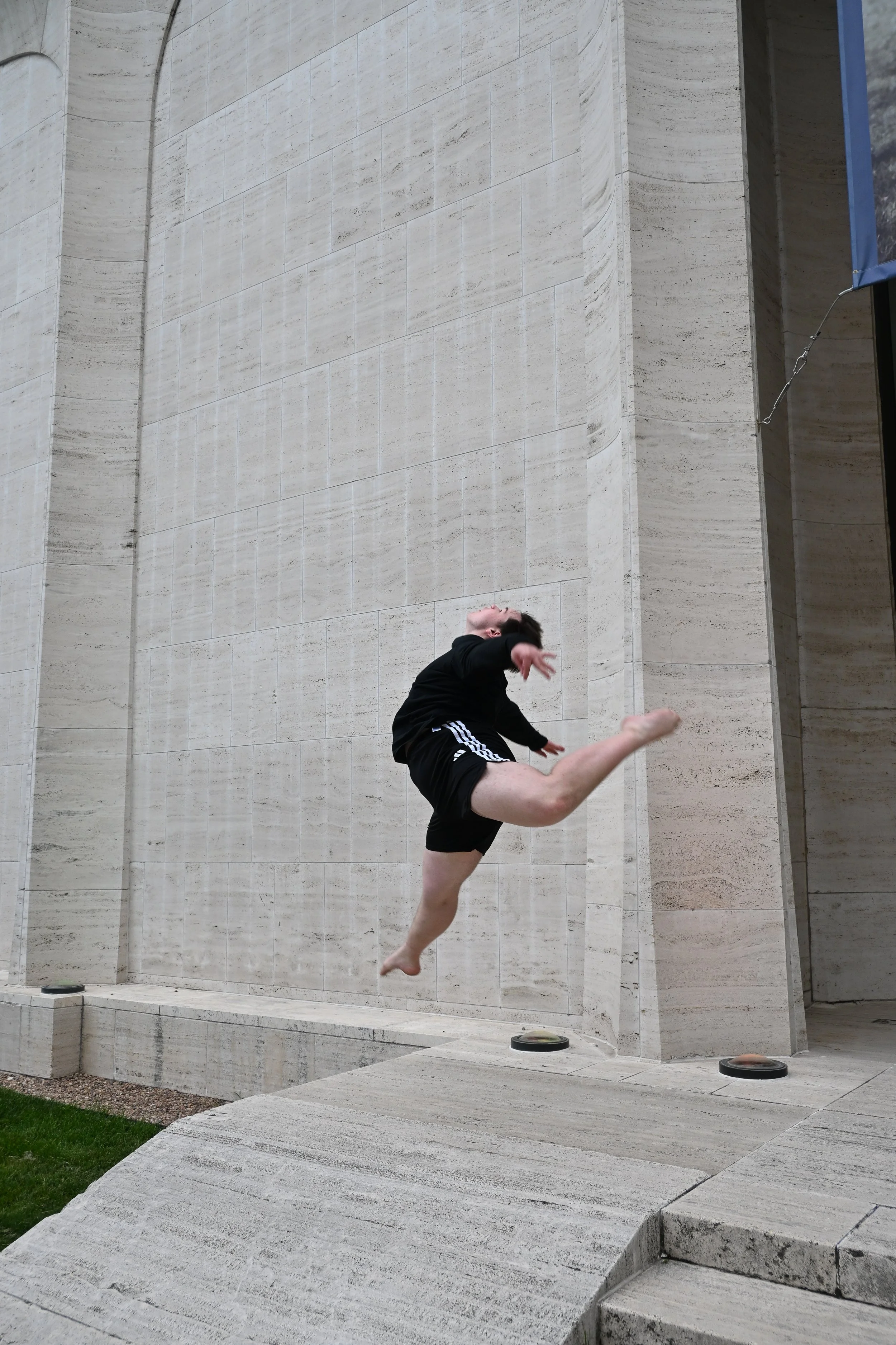A person mid-air performing a high kick or martial arts move in front of a large beige stone building with steps and a small grassy area.