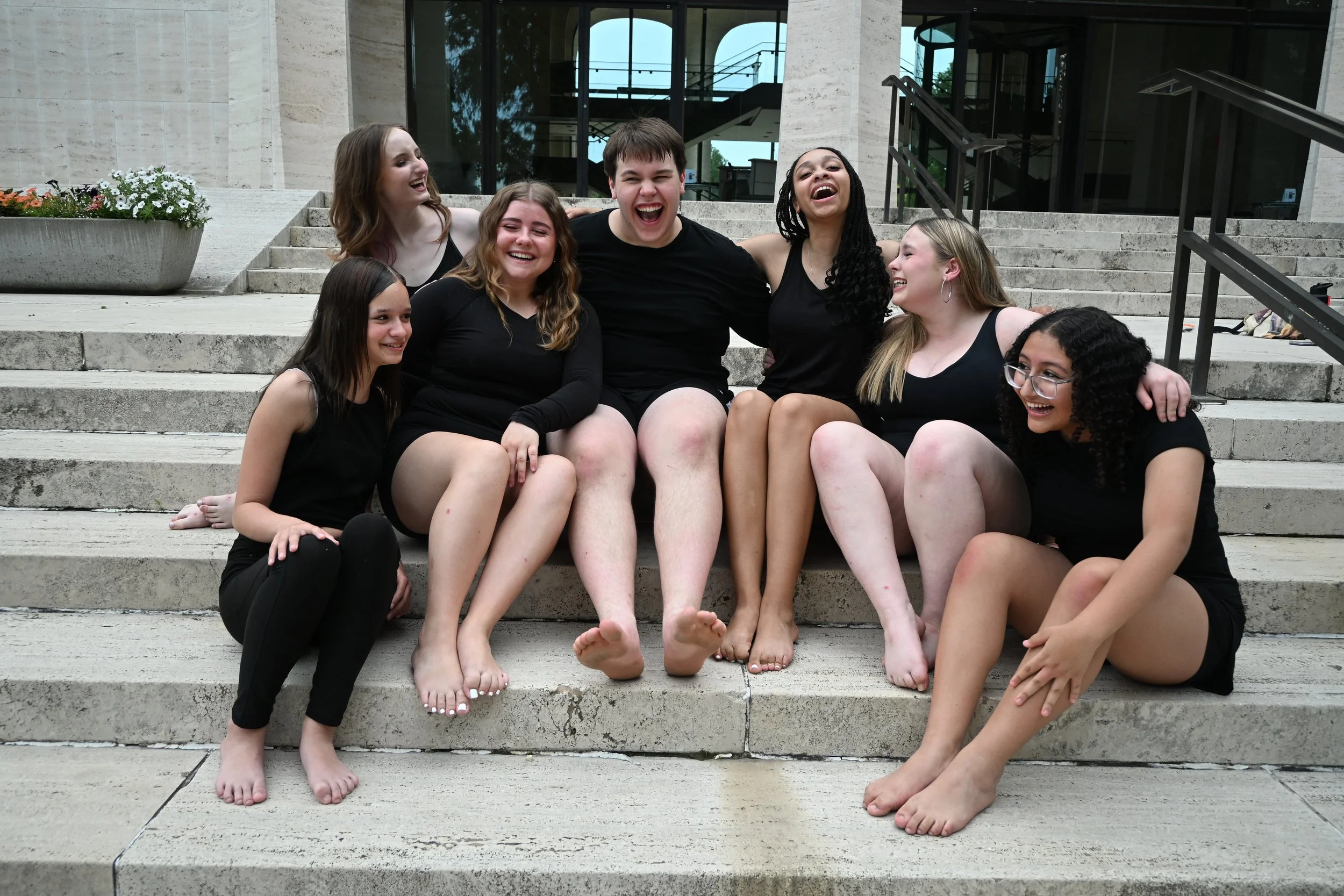 A group of seven young adults sitting on outdoor steps, wearing black clothing, smiling, and laughing together, with a modern building and glass windows in the background.