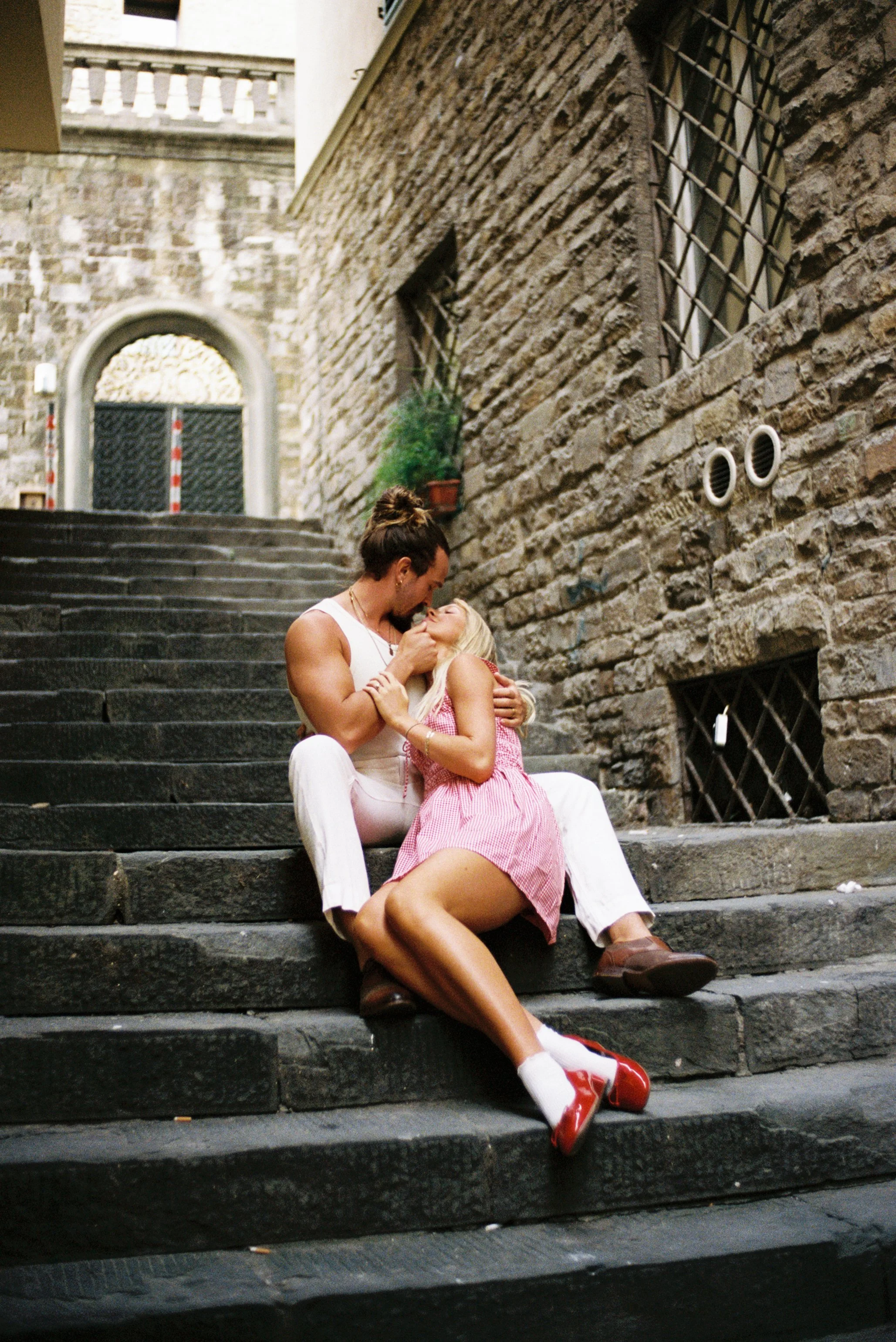 A couple sitting on stone stairs in an alleyway, sharing an intimate moment, with an old brick building in the background.