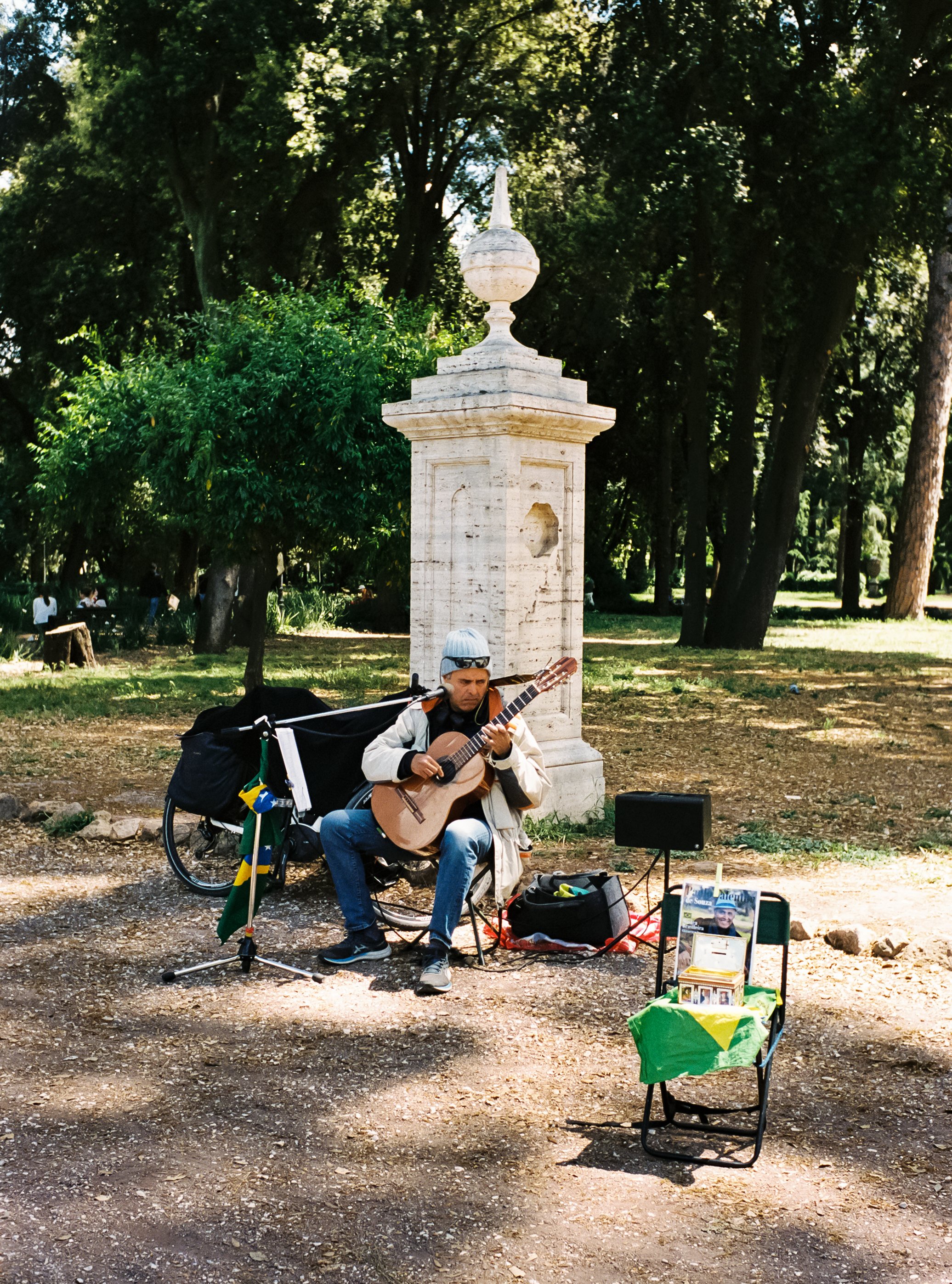 A street musician playing guitar outdoors in a park, with a small amplifier and a chair displaying pictures and donation items, with trees and a stone monument in the background.