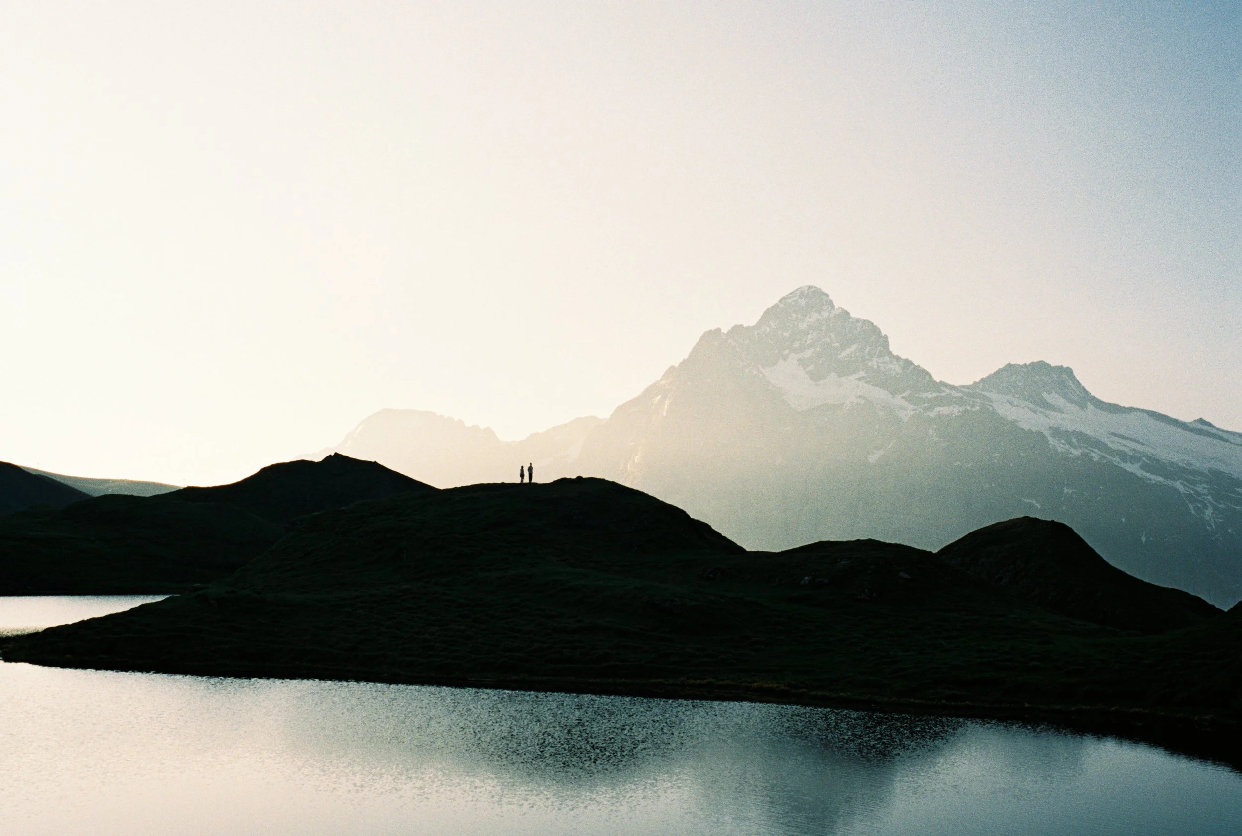 Silhouettes of two people standing on a grassy hilltop overlooking a mountain with snow, a lake in the foreground, and a clear sky.