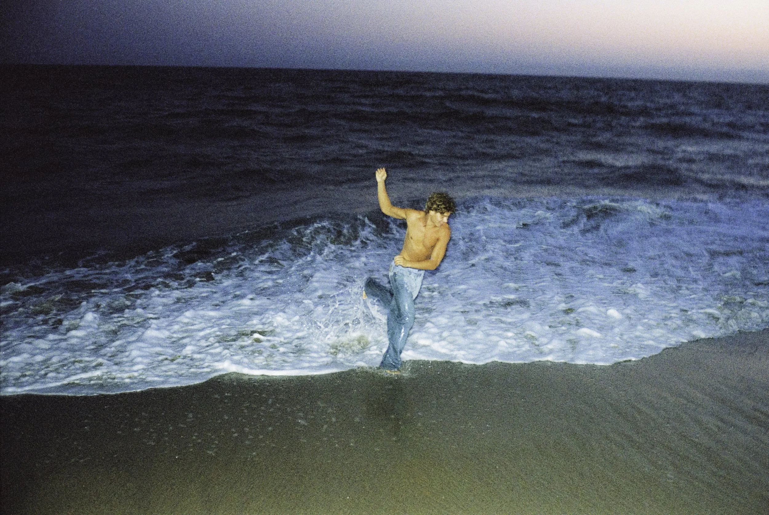 A shirtless man with curly hair wearing jeans is dancing or playing in the ocean waves near the shore at dusk or dawn.