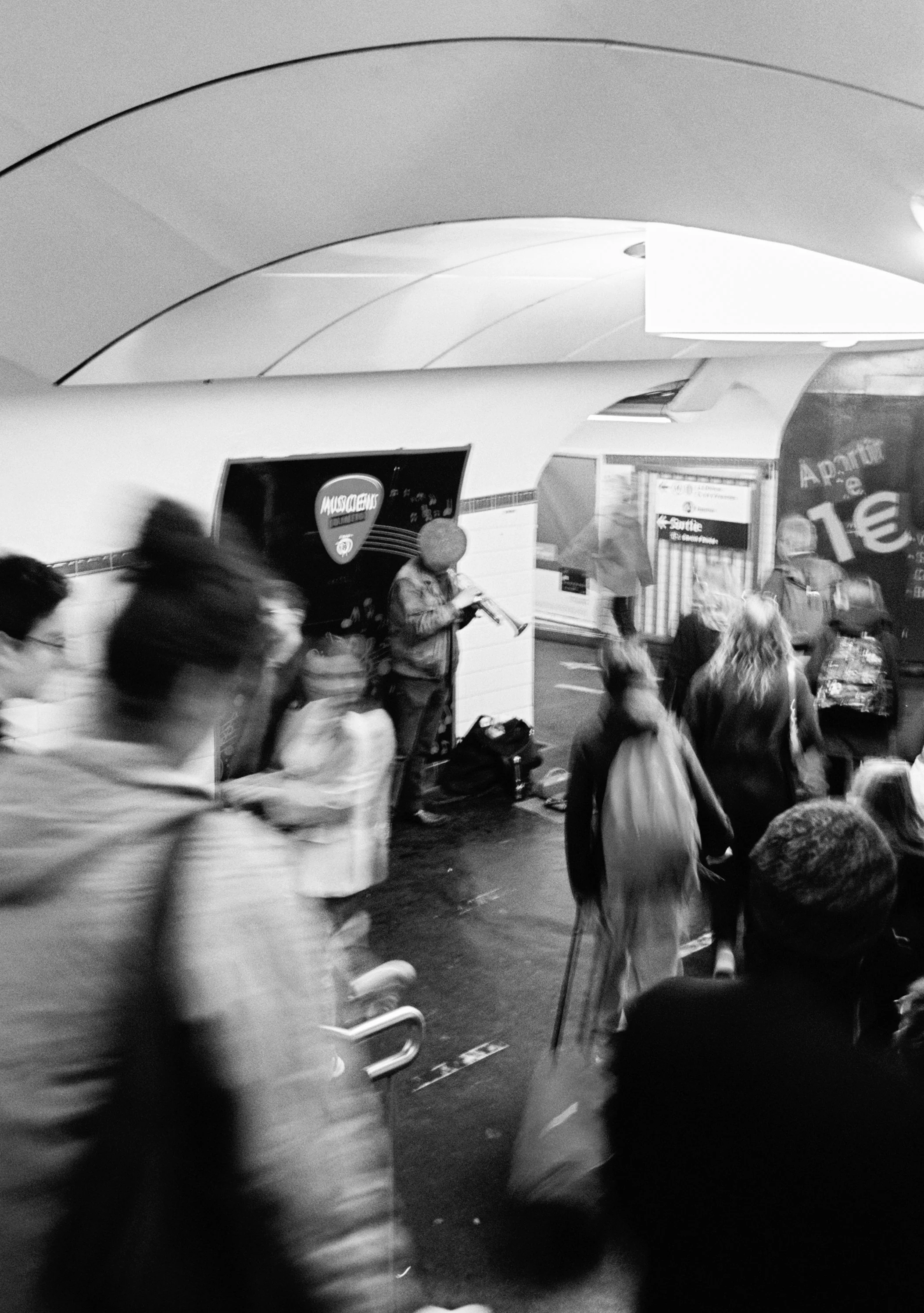 A black and white photo of an underground station with a live street musician playing a violin, surrounded by a blurred crowd of people walking past.