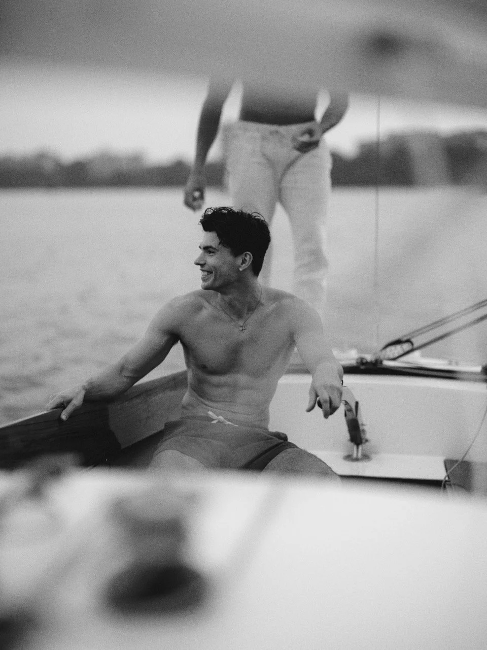 A young man smiling and sitting shirtless on a boat, with another person standing behind him in a boat on the water.