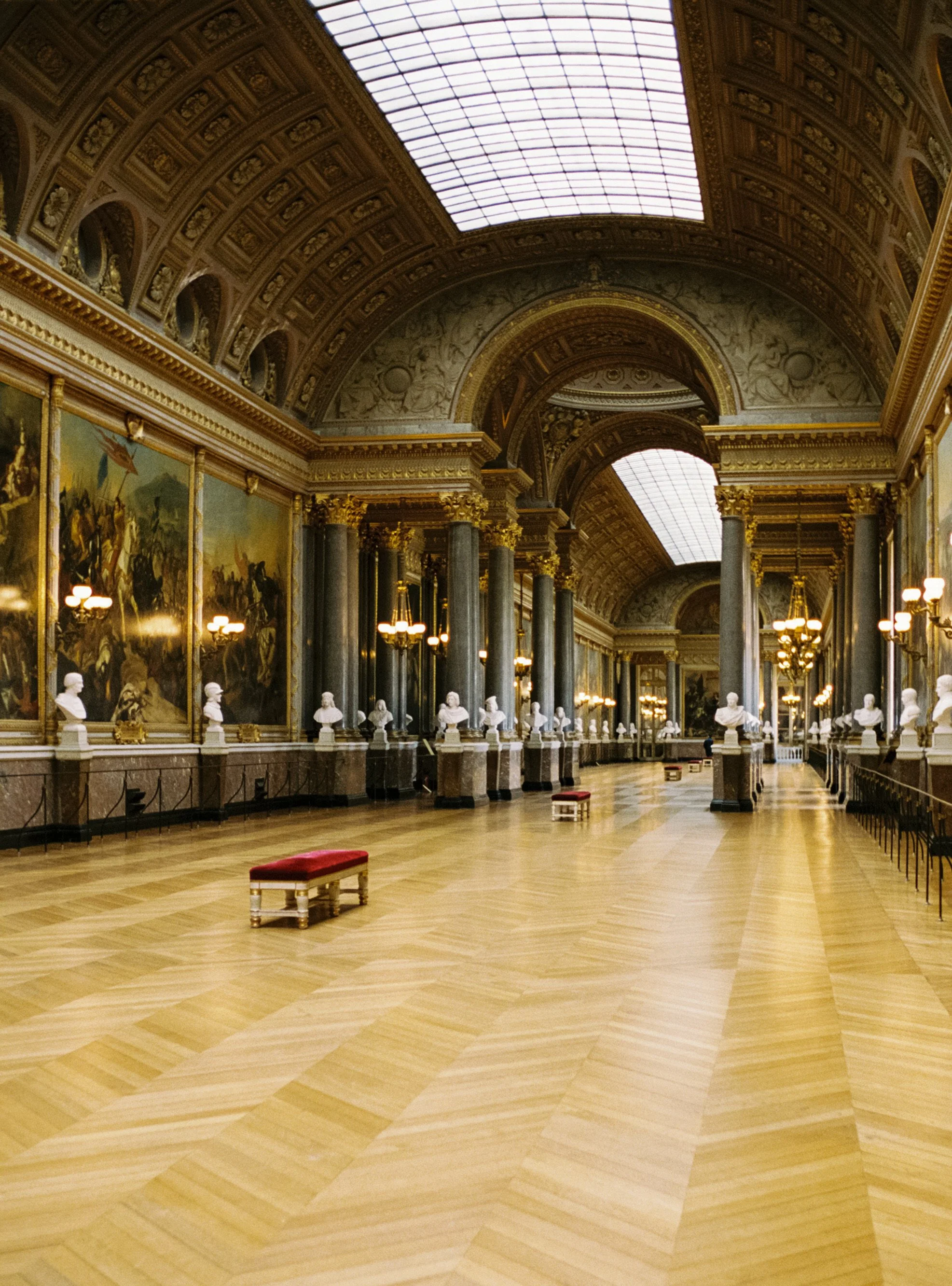 A grand, ornately decorated hall with high ceilings, large skylights, marble columns, and portraits or paintings on the walls. There are bust sculptures on pedestals and small red velvet benches along the floor.