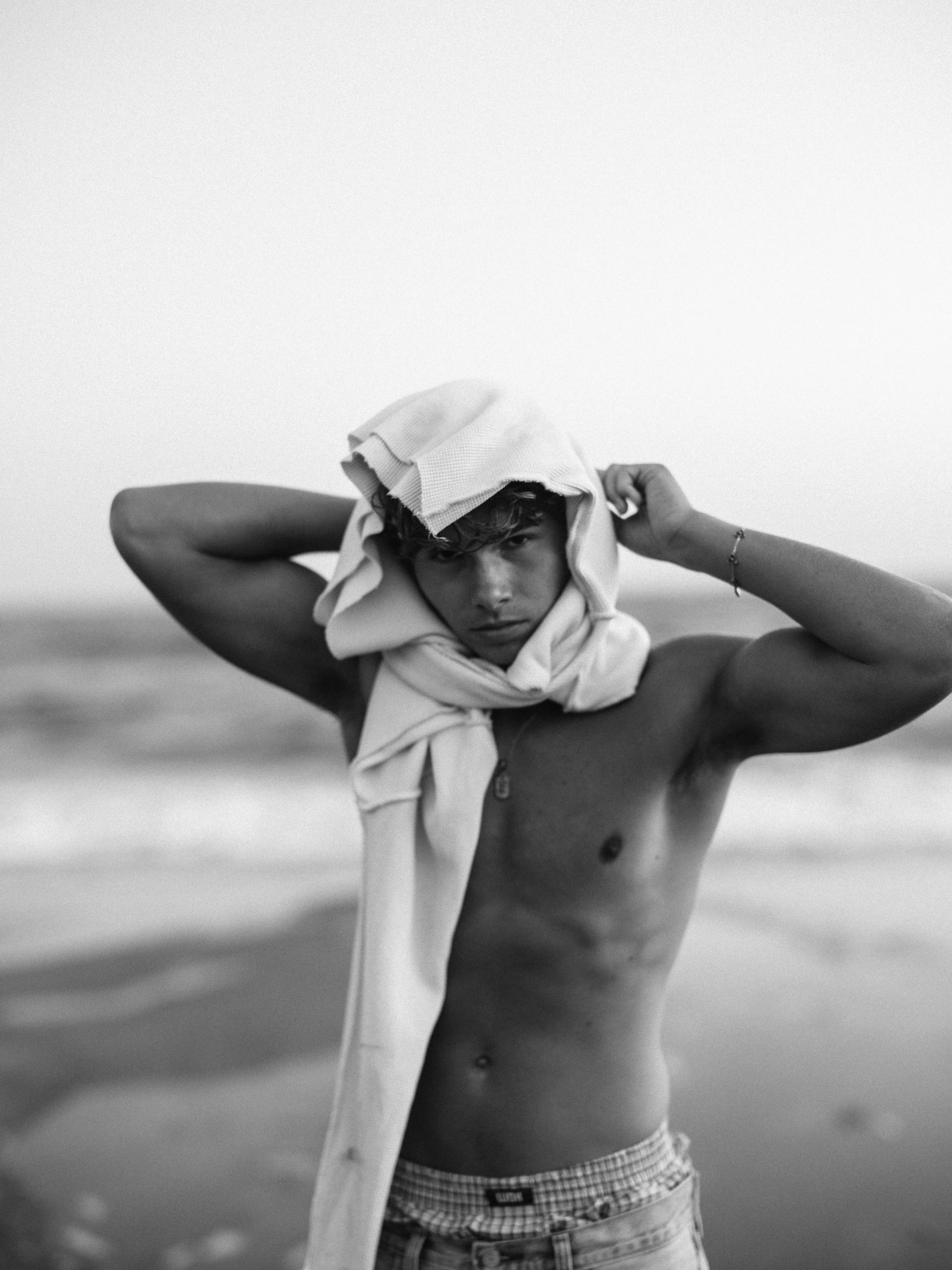 A shirtless young man stands on the beach with a towel draped over his head, looking directly at the camera with a serious expression.