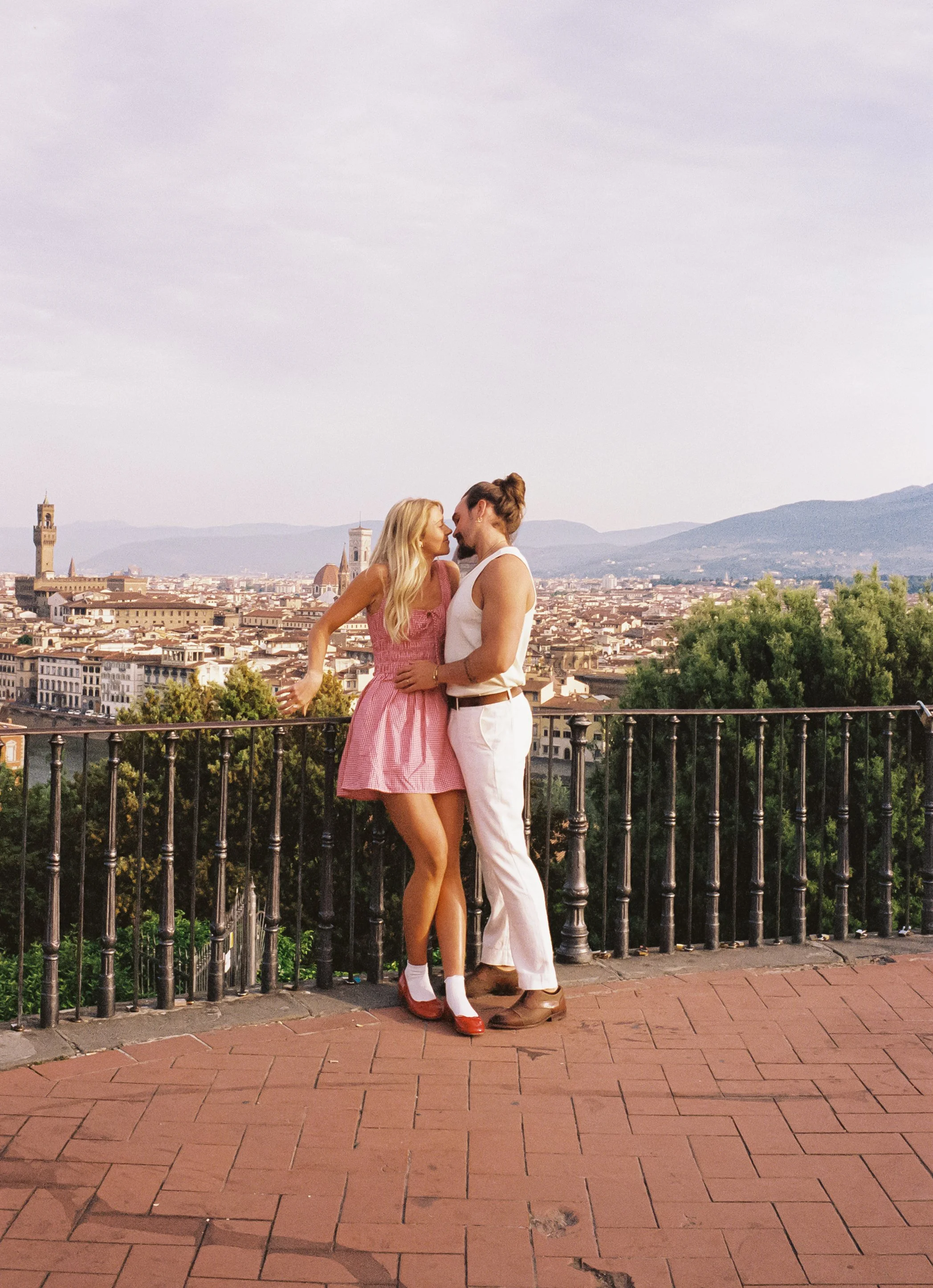 A couple standing close and gazing at each other on a terrace with a cityscape background, including historic buildings and mountains.