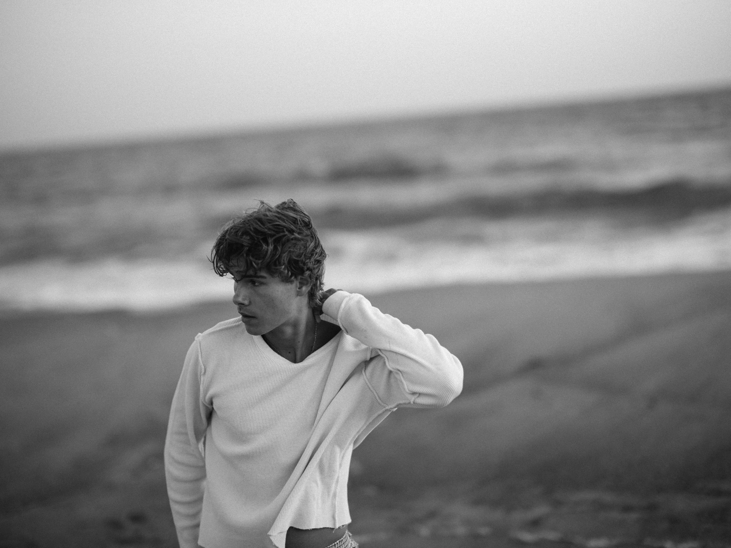 A young man with wavy hair stands on a beach with waves in the background, wearing a light-colored long-sleeve shirt, looking to the side with a contemplative expression.