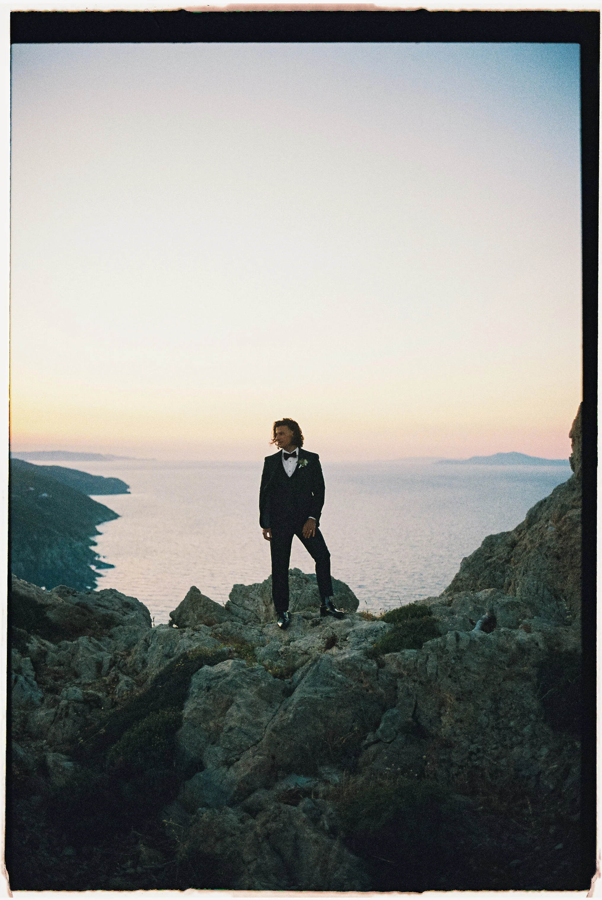 A man in a tuxedo standing on rocks near the ocean during sunset.
