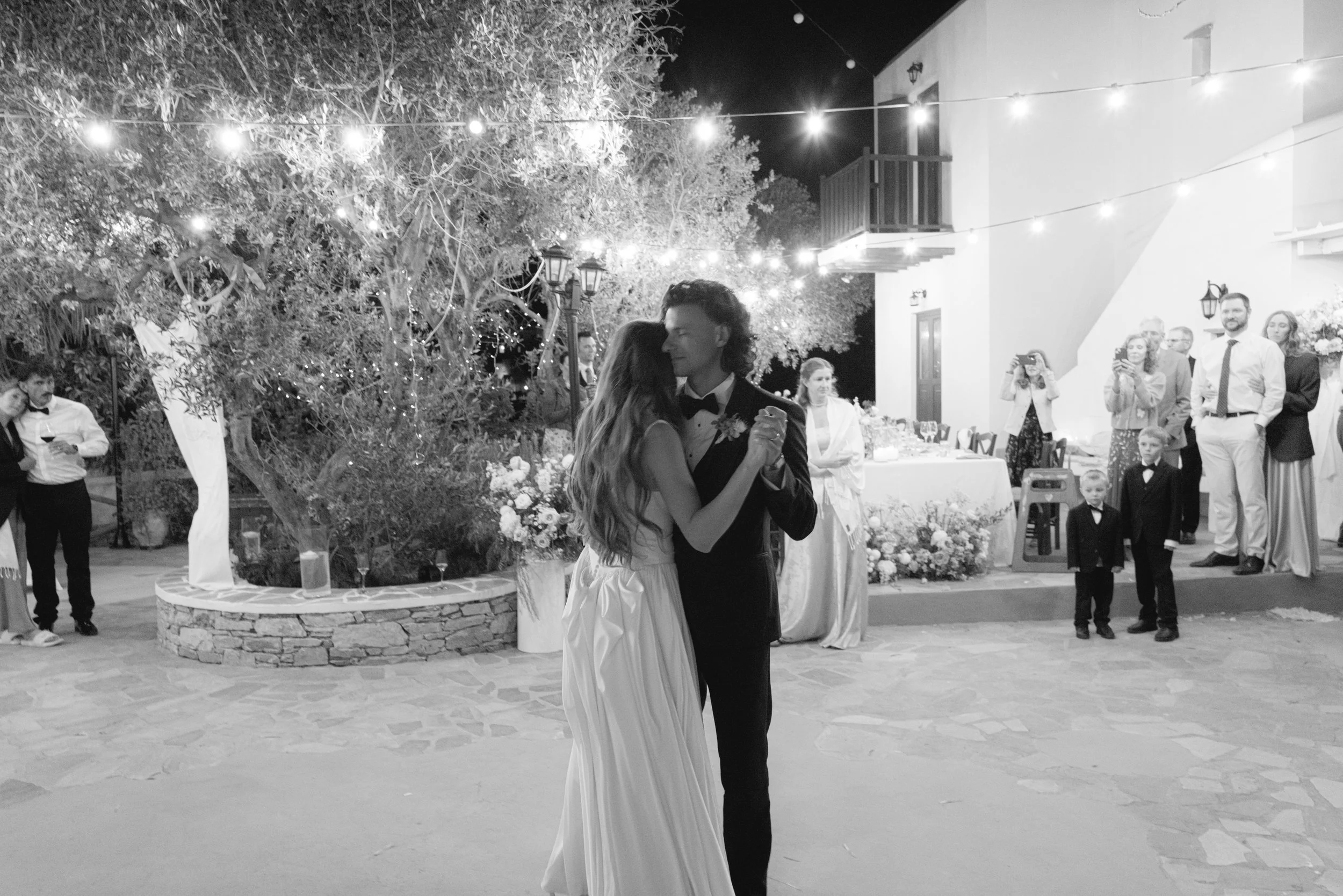 A bride and groom dancing at their wedding reception outdoors at night, with guests watching and taking pictures, string lights hanging overhead, and festive decorations around.