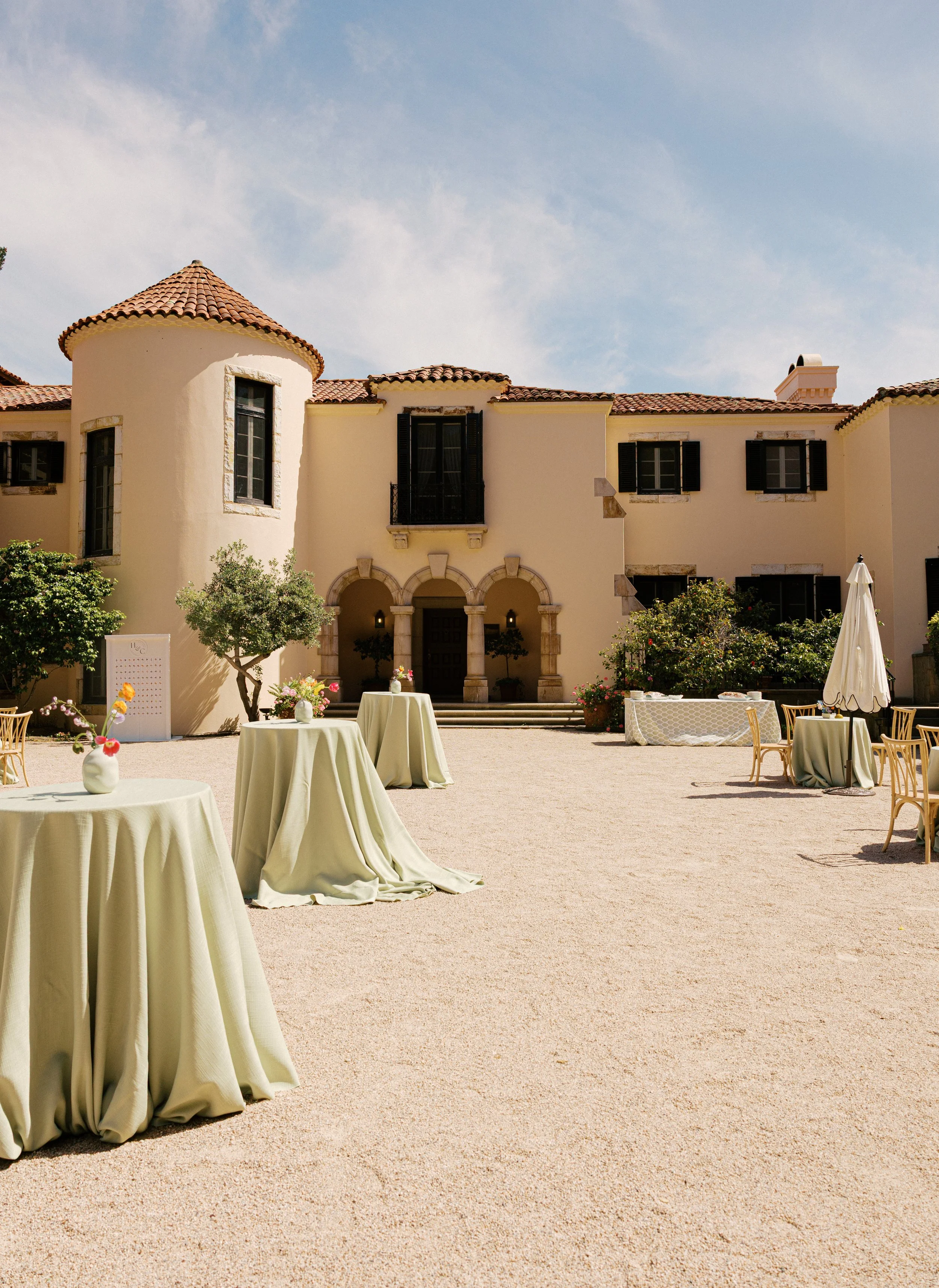 An outdoor courtyard with several tables covered in beige tablecloths and decorated with vases of flowers, set in front of a large, elegant cream-colored building with a tiled roof, arched entryways, and black window shutters. The sky is partly cloud