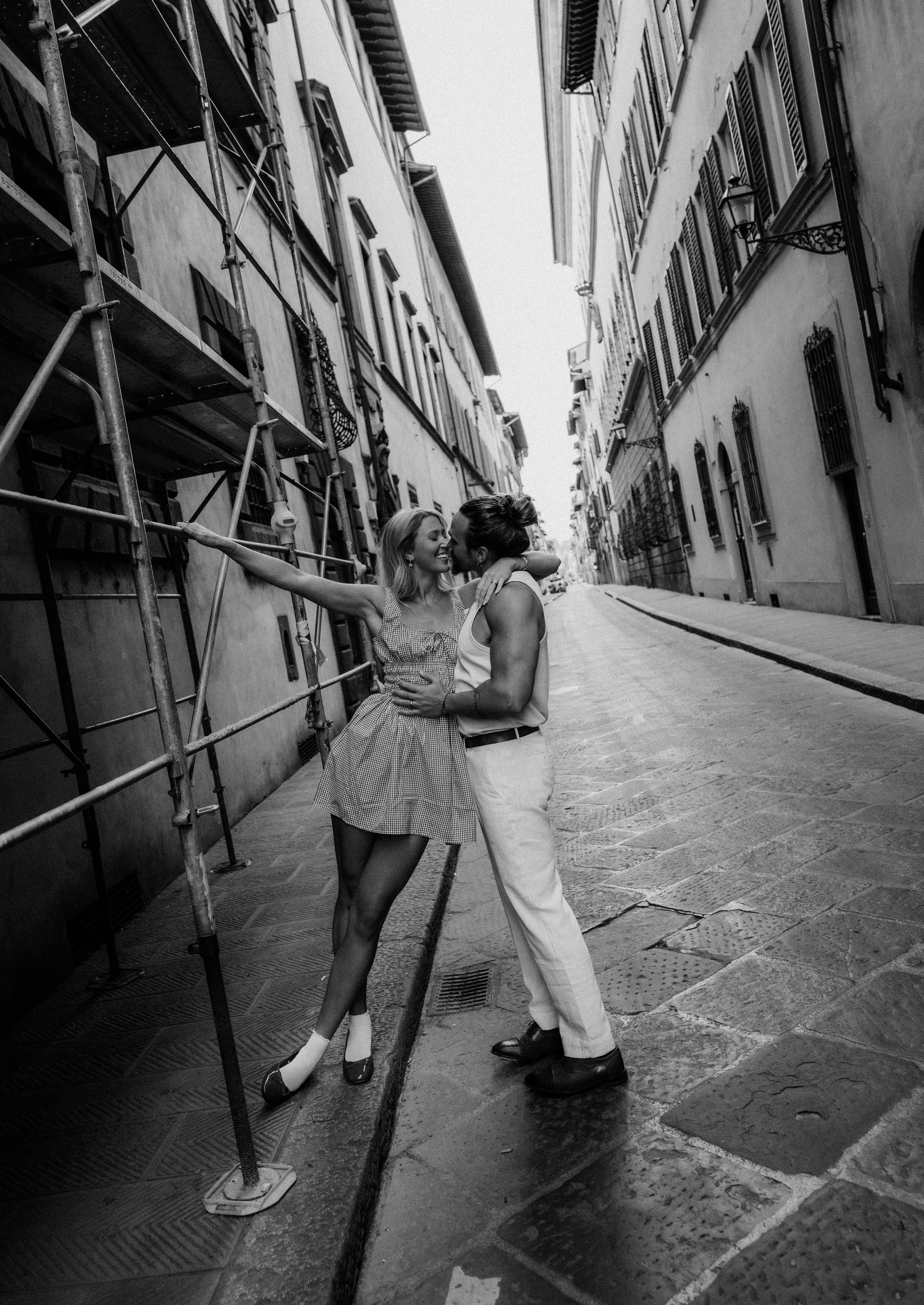Two women embrace and smile at each other in the middle of a narrow street with old buildings on either side. One woman is wearing a gingham dress and high heels, while the other is dressed in light-colored pants and a sleeveless top. The scene is in