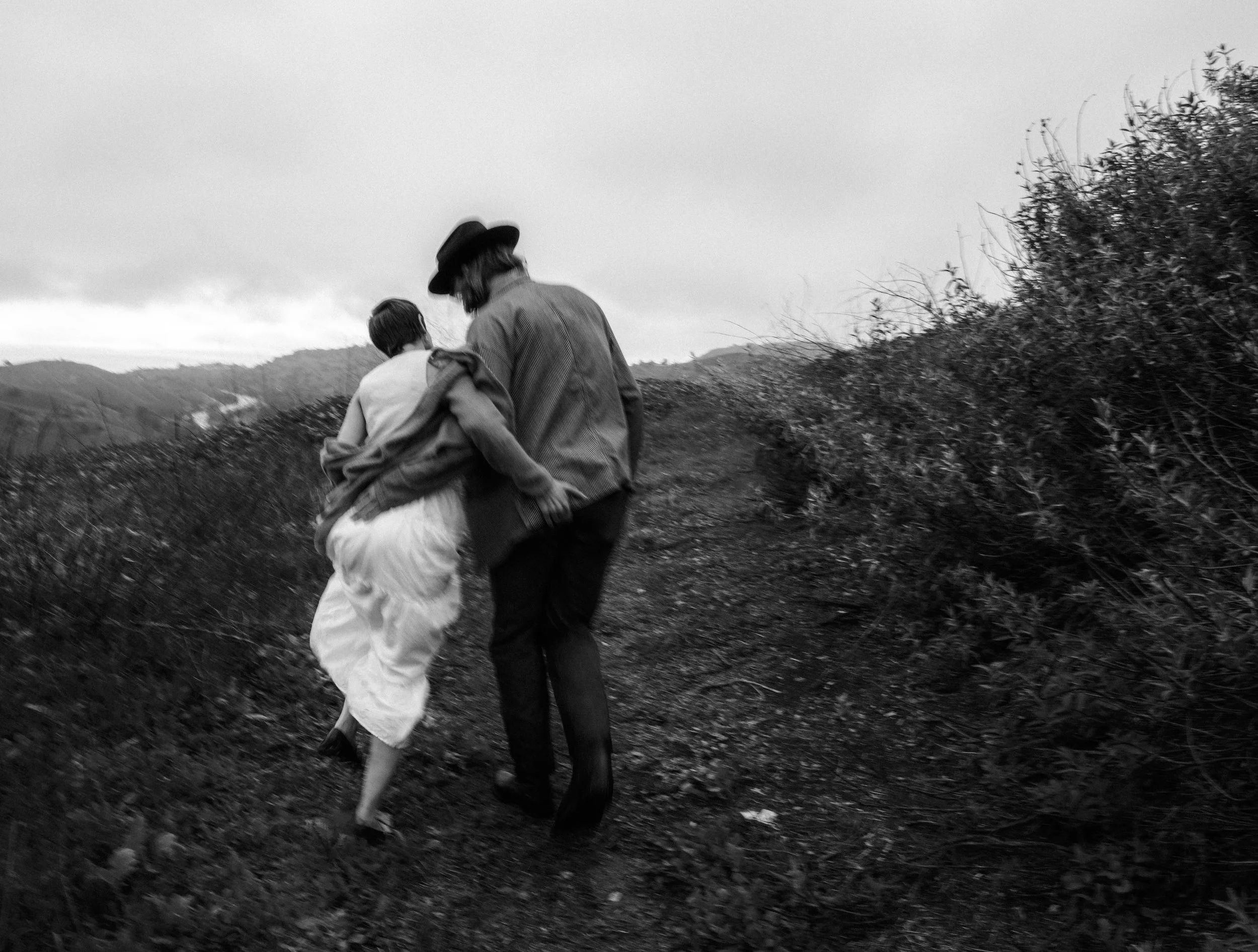 Black and white photo of two people walking up a dirt path with bushes on either side, hills in the background, one person wearing a hat, both dressed casually.