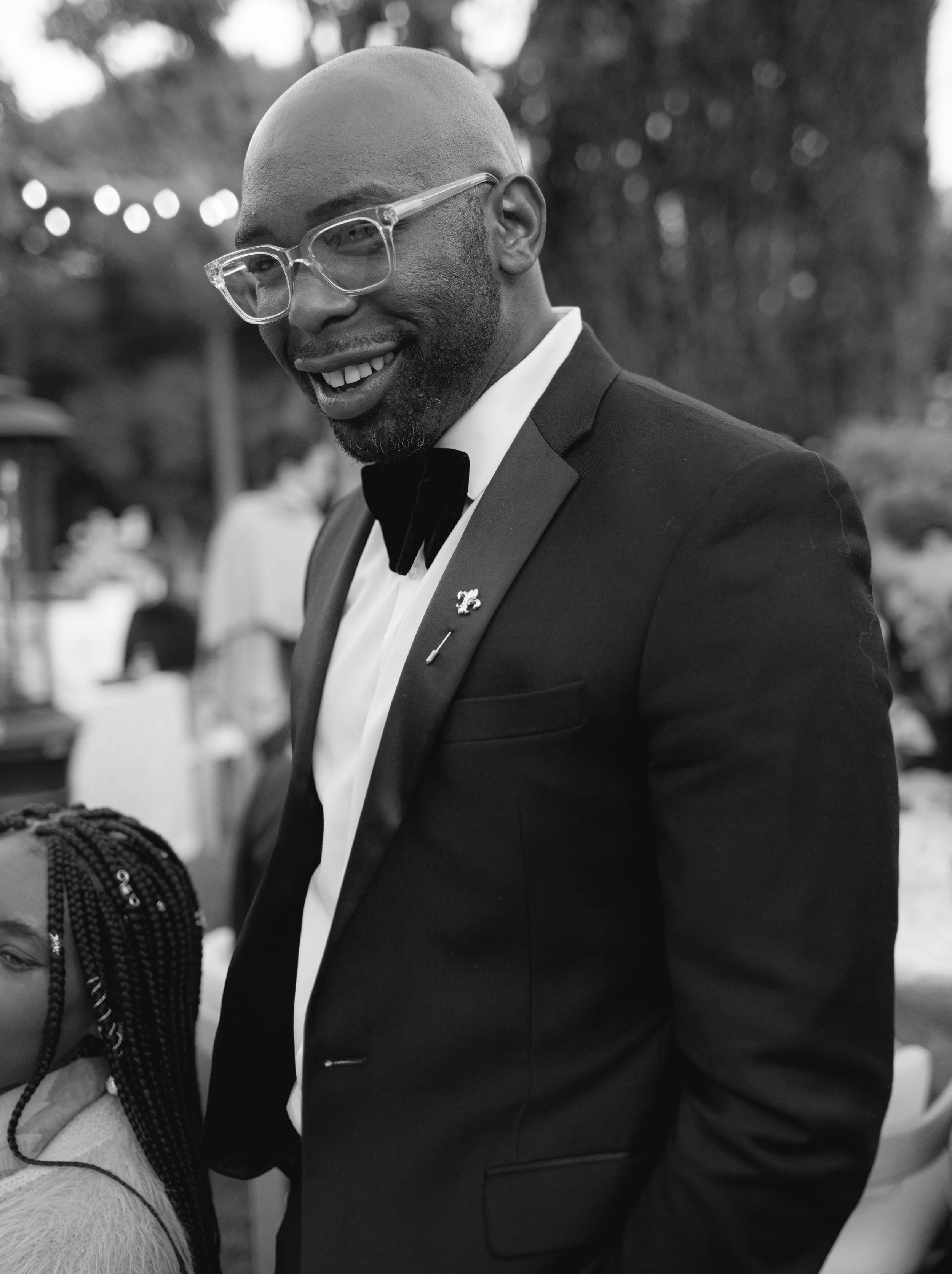 Black and white photo of a smiling, bald man wearing glasses and a tuxedo with a small flower pin, outdoors with blurred trees and lights in the background.