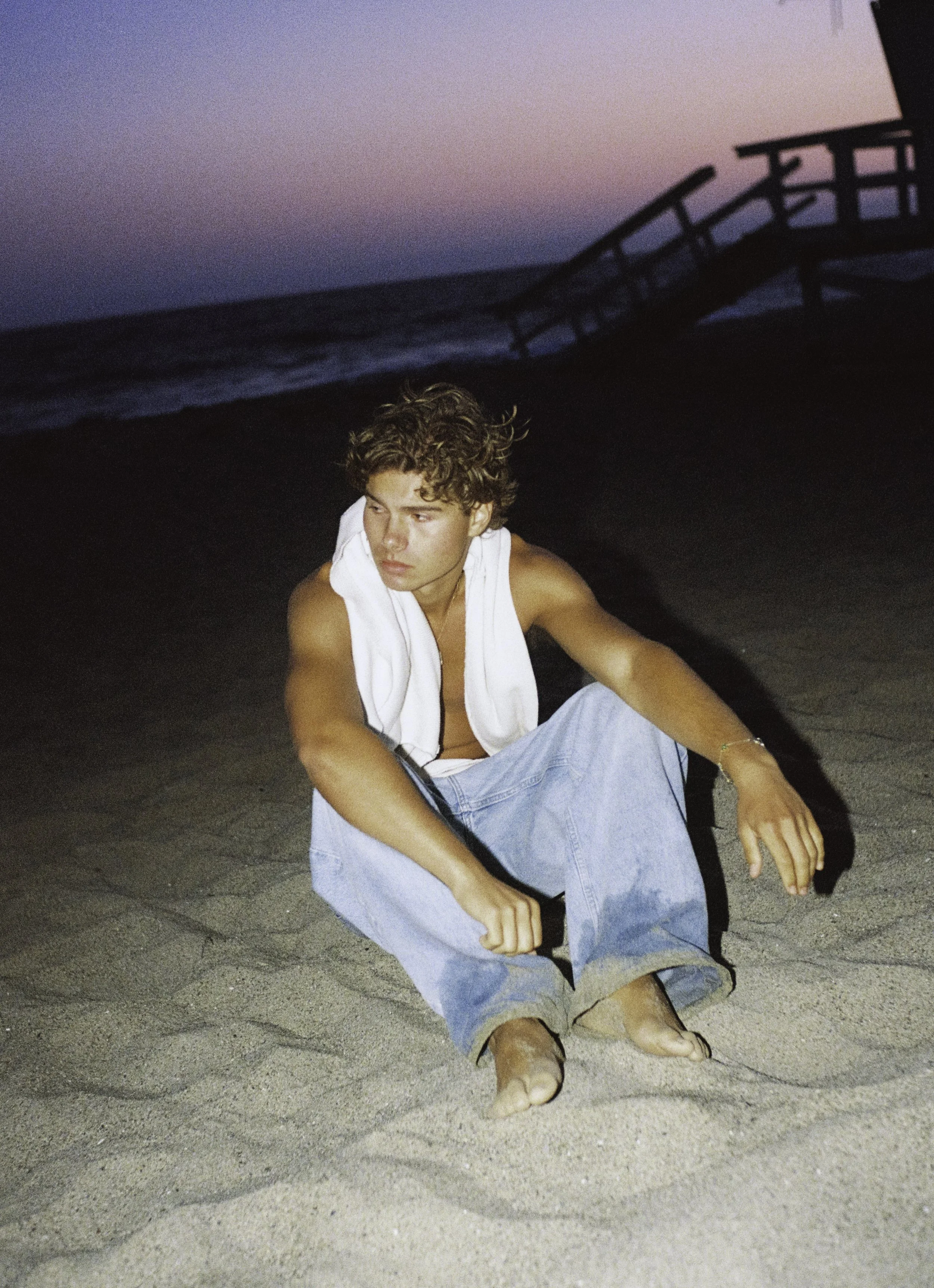 A young man with curly hair sits on the sandy beach at dusk, wearing baggy jeans and a sleeveless shirt, with a towel draped over his shoulders, and an ocean and sunset in the background.