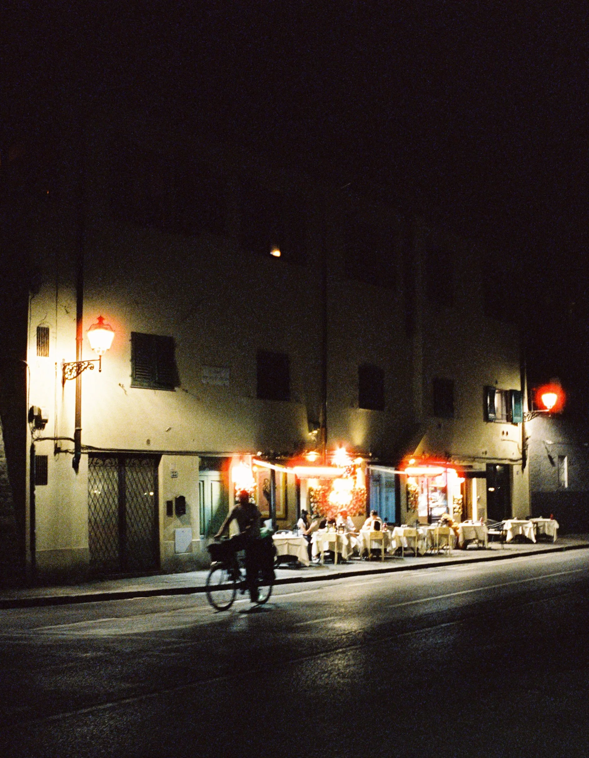 Nighttime scene of a restaurant with outdoor seating, illuminated by warm lights, with a cyclist passing by on the street in the foreground.