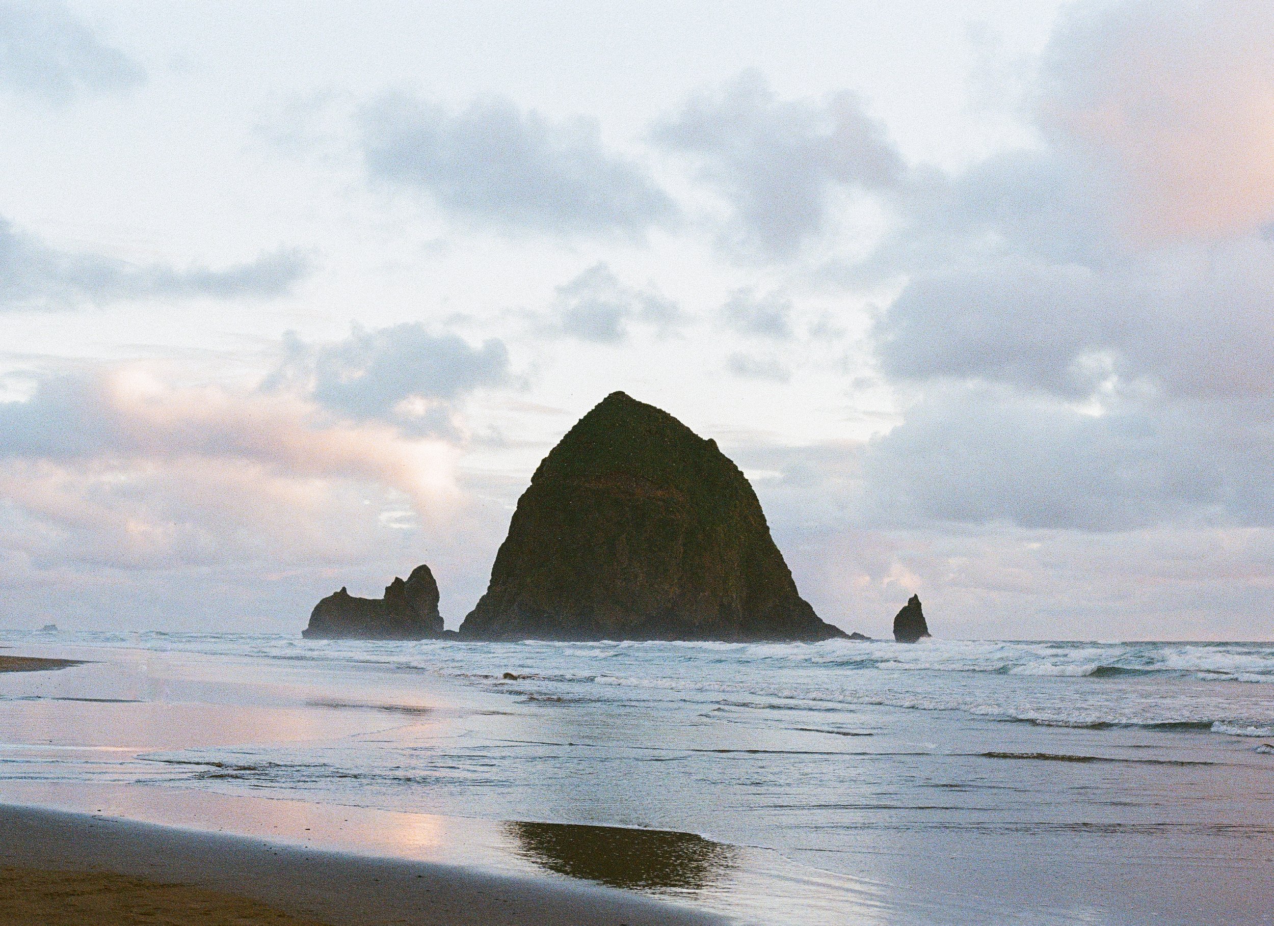 Ocean waves crashing on a sandy beach with large rock formations in the background under a cloudy sky.