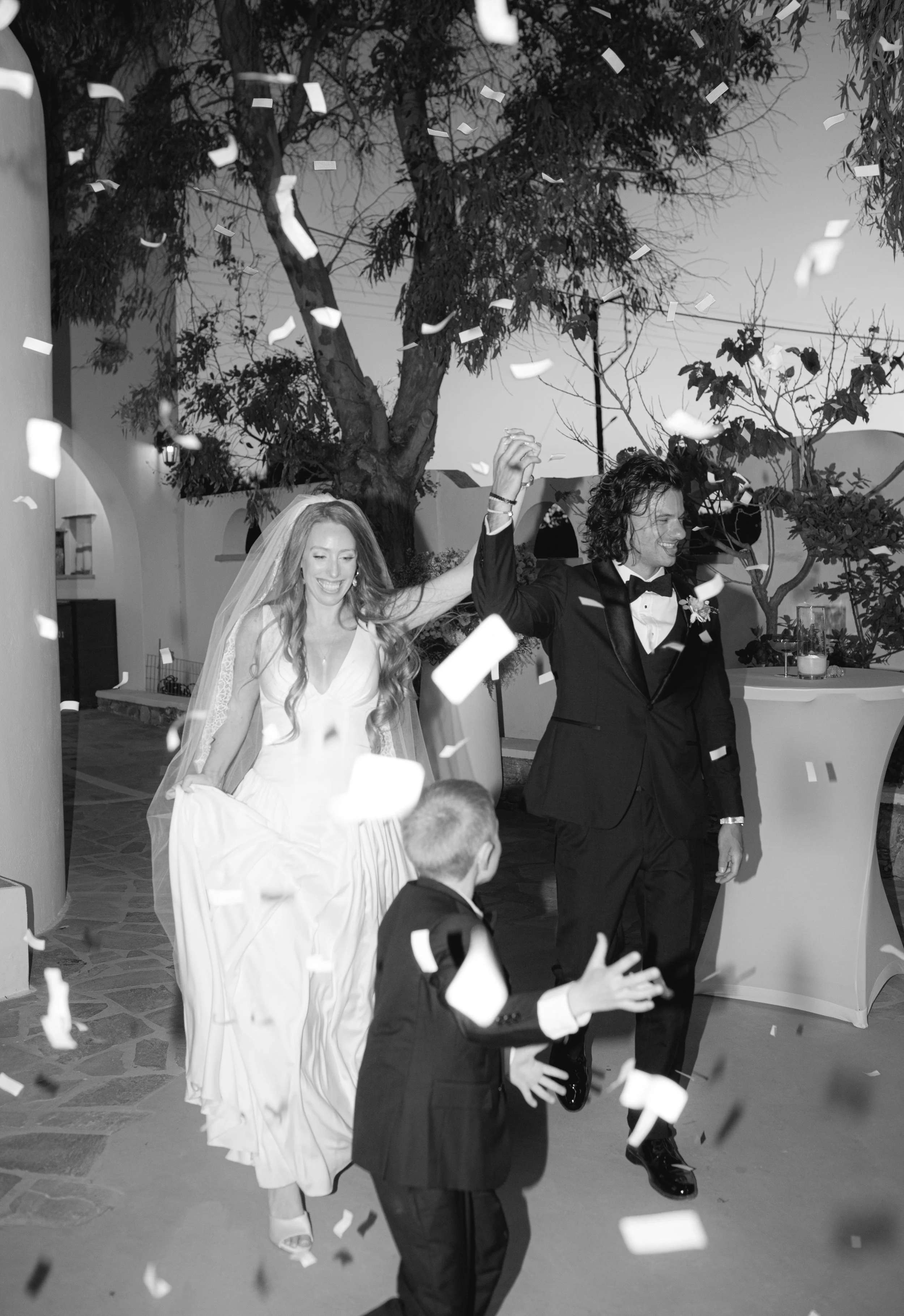 A bride and groom celebrating wedding reception with confetti falling around them. The bride wears a wedding dress and veil, smiling, and the groom in a tuxedo, also smiling and holding hands up.