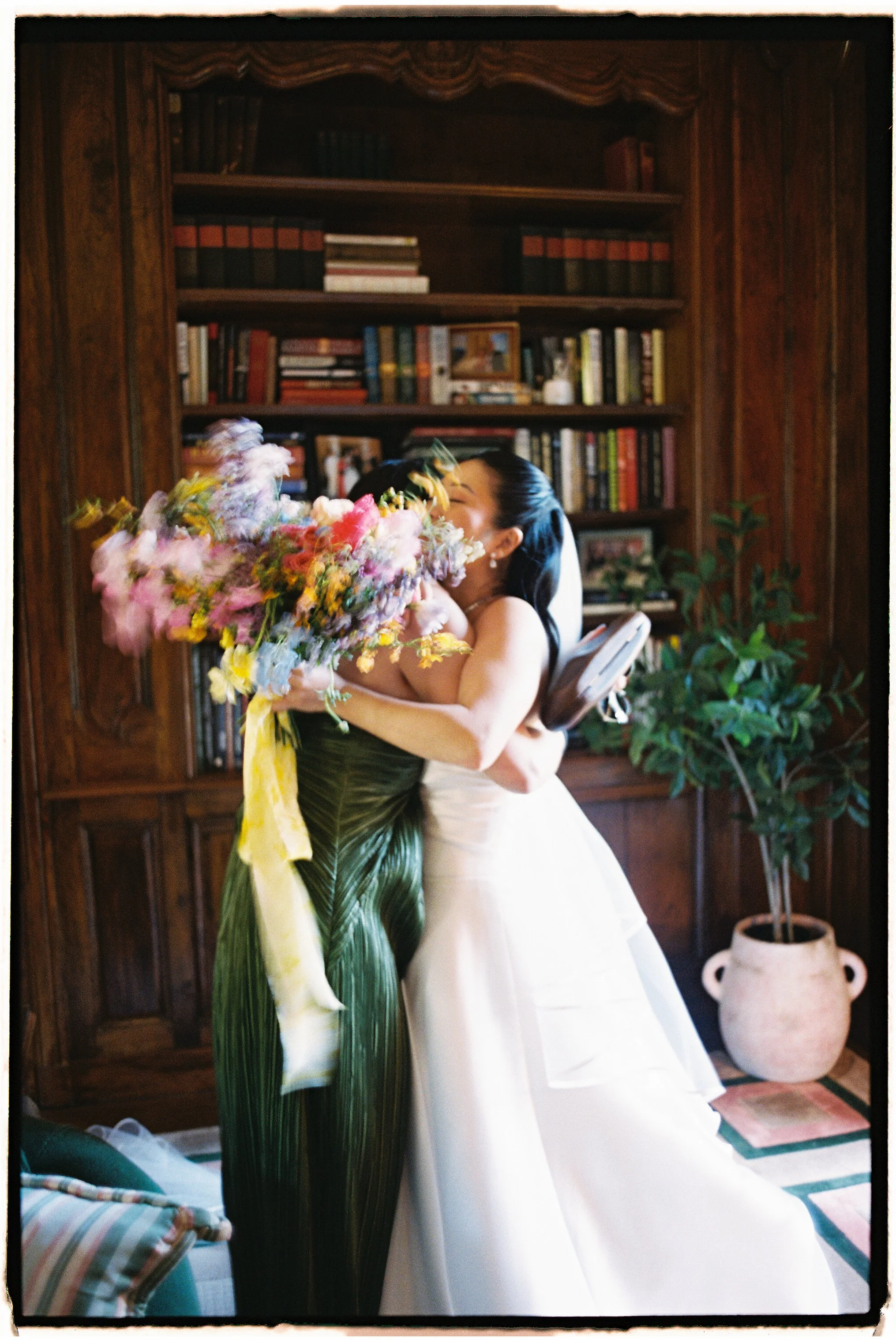 A woman in a white wedding dress hugging another woman in a green leaf-patterned dress, holding a large bouquet of flowers, in a room with wooden bookshelves and a potted plant.