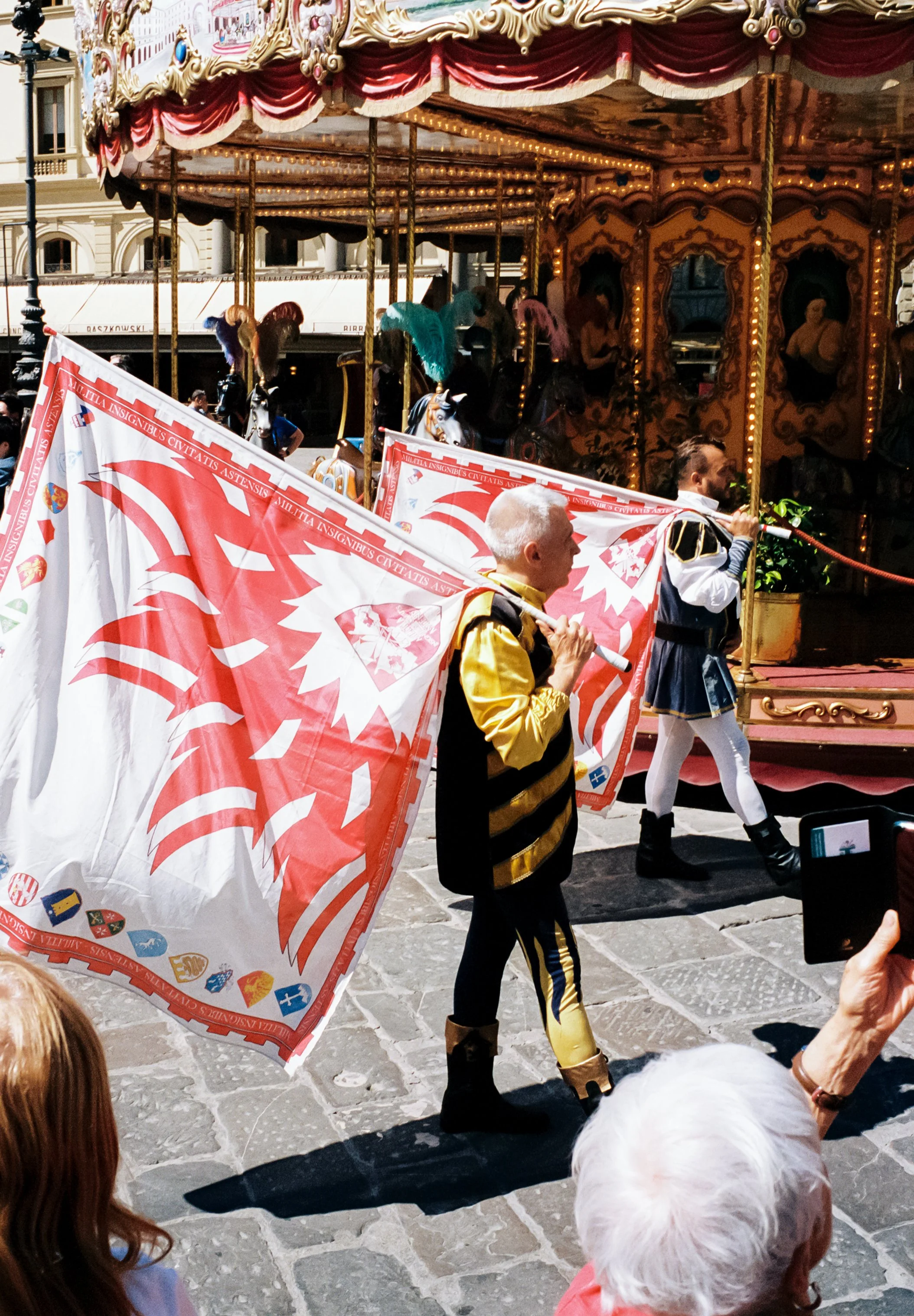 People participating in a parade dressed in historical costumes, carrying a large flag with a red and white lion emblem, walking past a ornate, gold-trimmed carousel in a European city square.