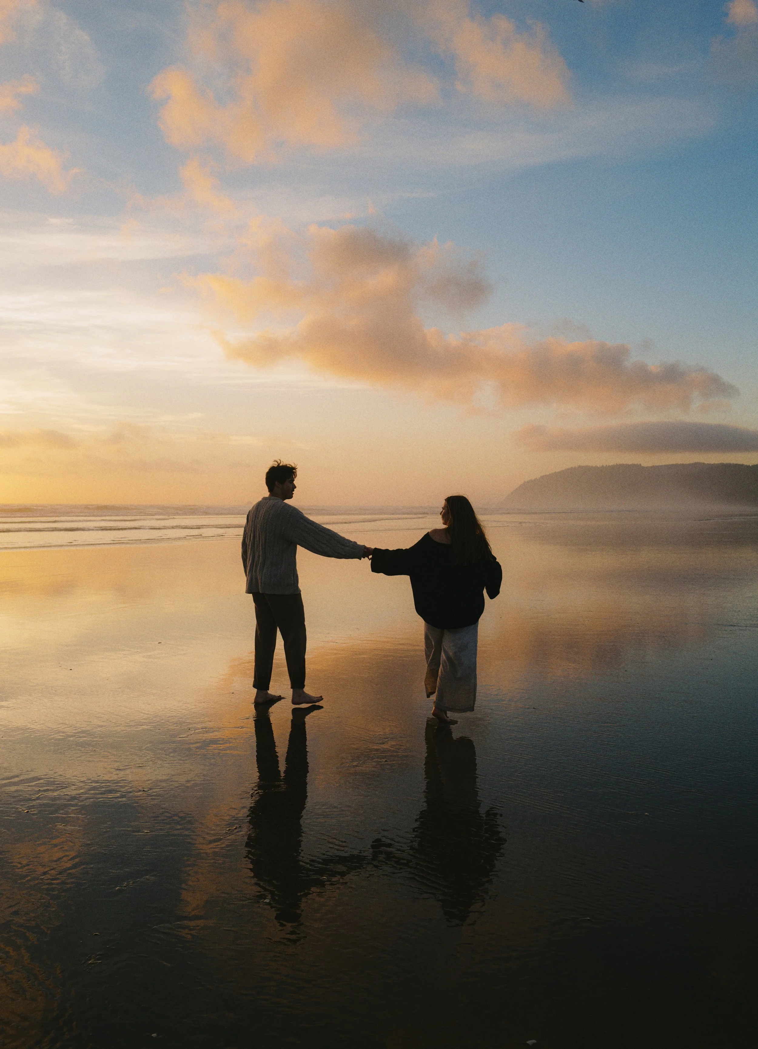 a girl and a boy holding hands walking into the ocean at sunset and golden hour