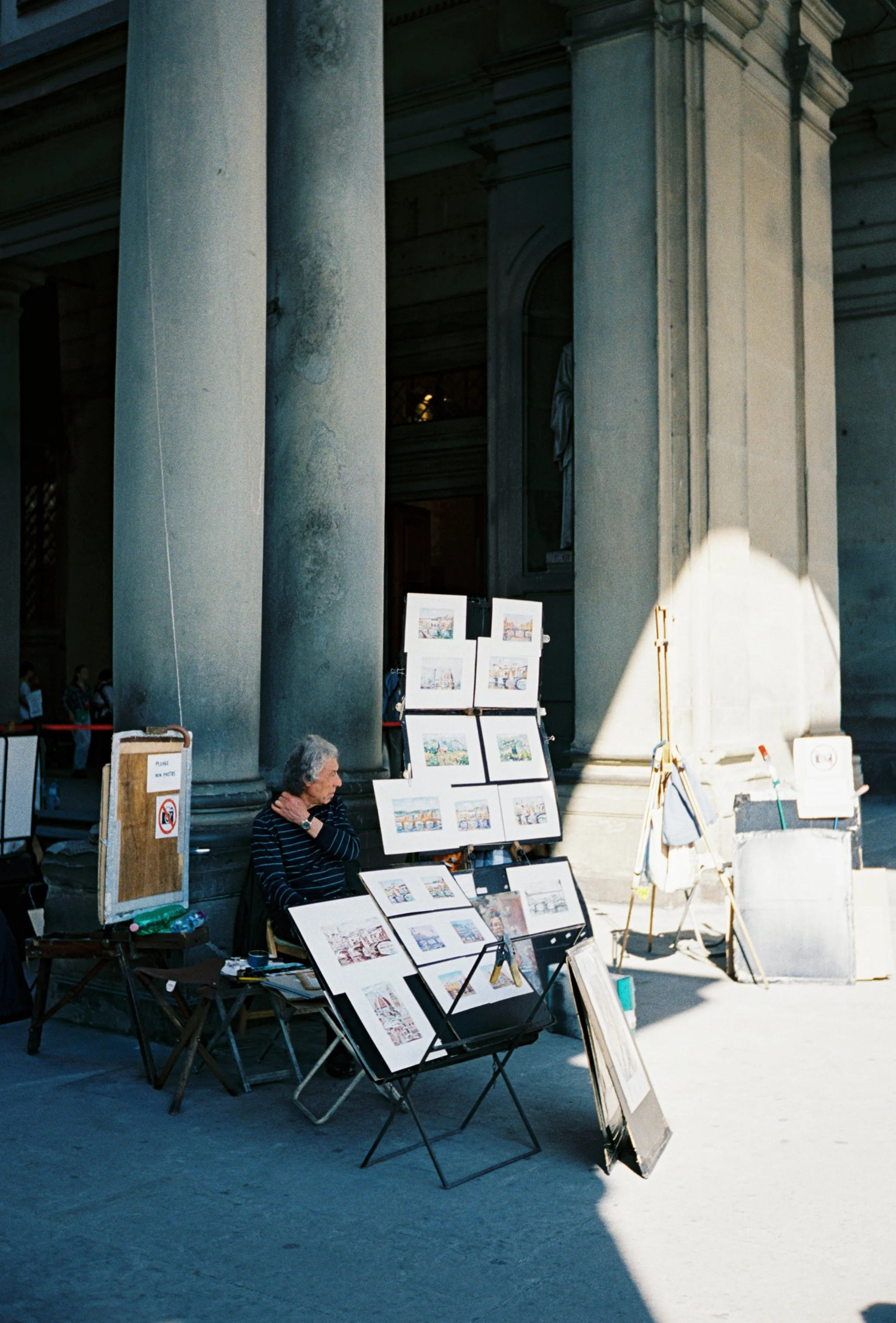 Street artist selling watercolor paintings of cityscapes outside a historic building with large columns.