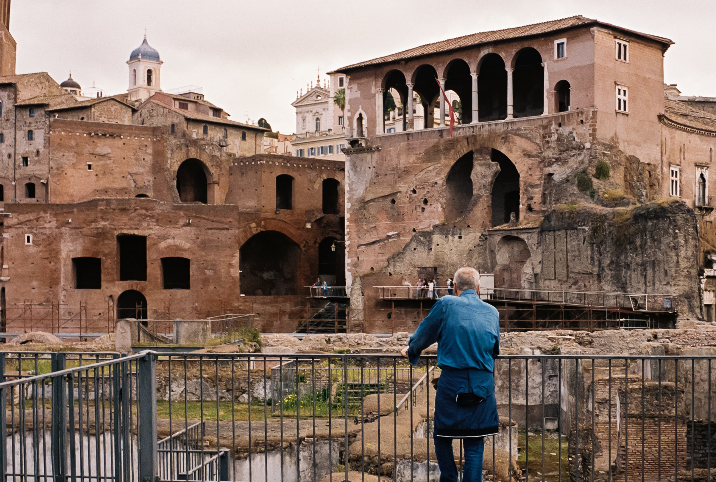 An elderly man in a blue jacket standing behind a metal railing, looking at ancient ruins with multi-level stone and brick structures, arches, and windows, and a few tourists exploring the site, likely in Rome, Italy.