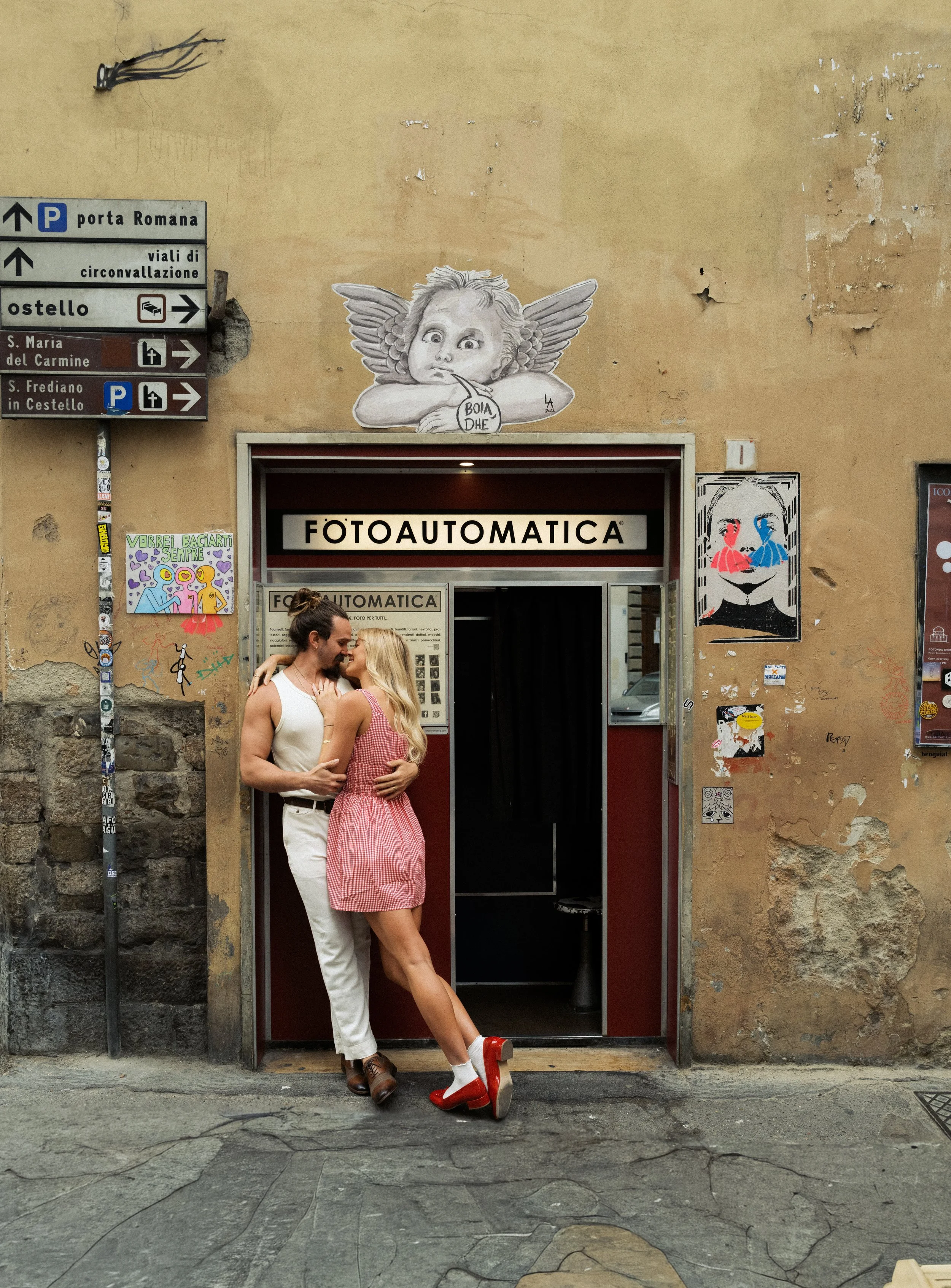 Two people, a man and a woman, are standing close together in front of the entrance to a photo booth called 'FOTOAUTOMATICA'. The woman is wearing a pink and white checkered dress and red high heel shoes, while the man is dressed in a white outfit an