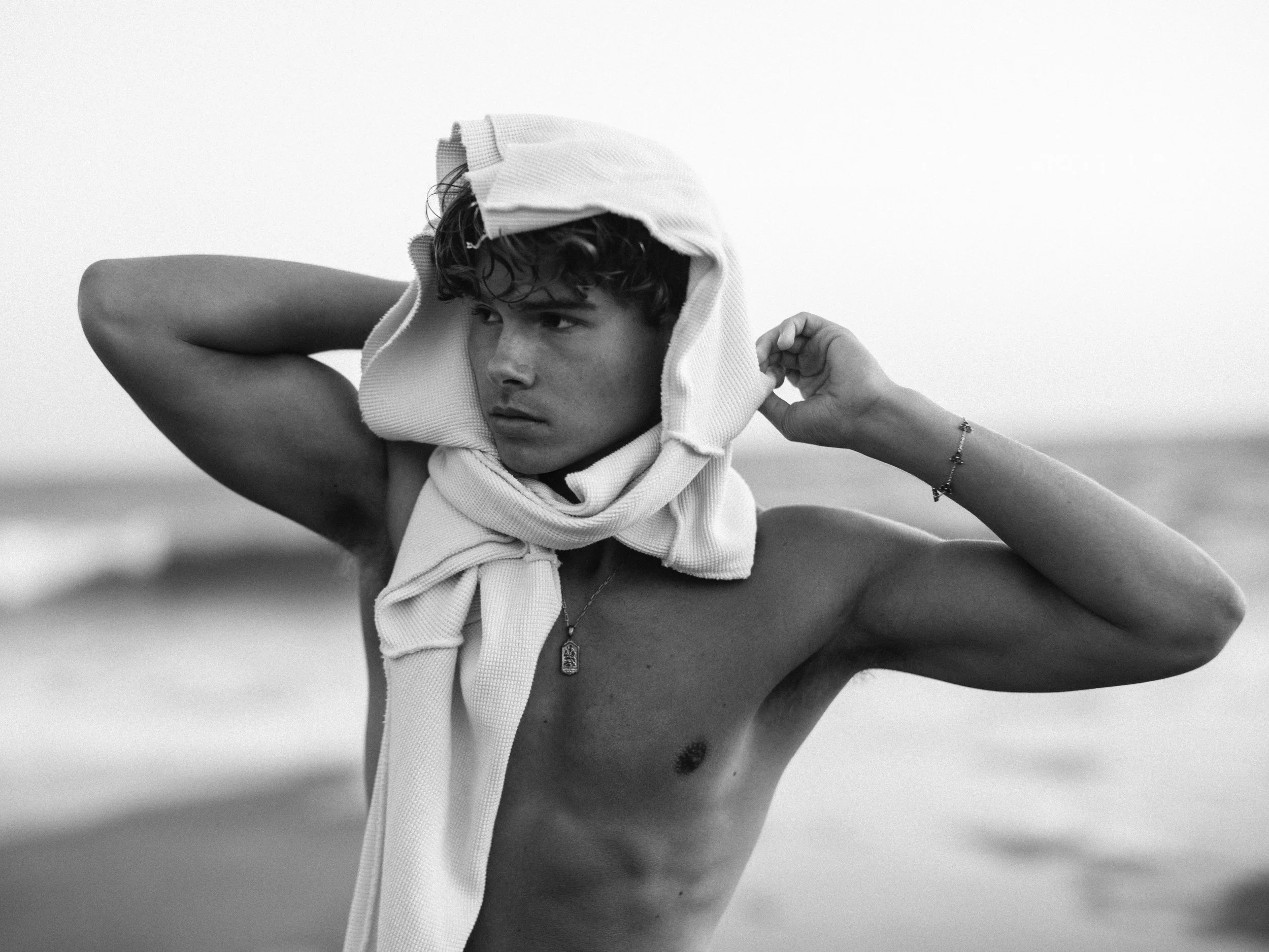Black and white photo of a young shirtless man on the beach, adjusting a towel around his head, with a necklace and bracelet.