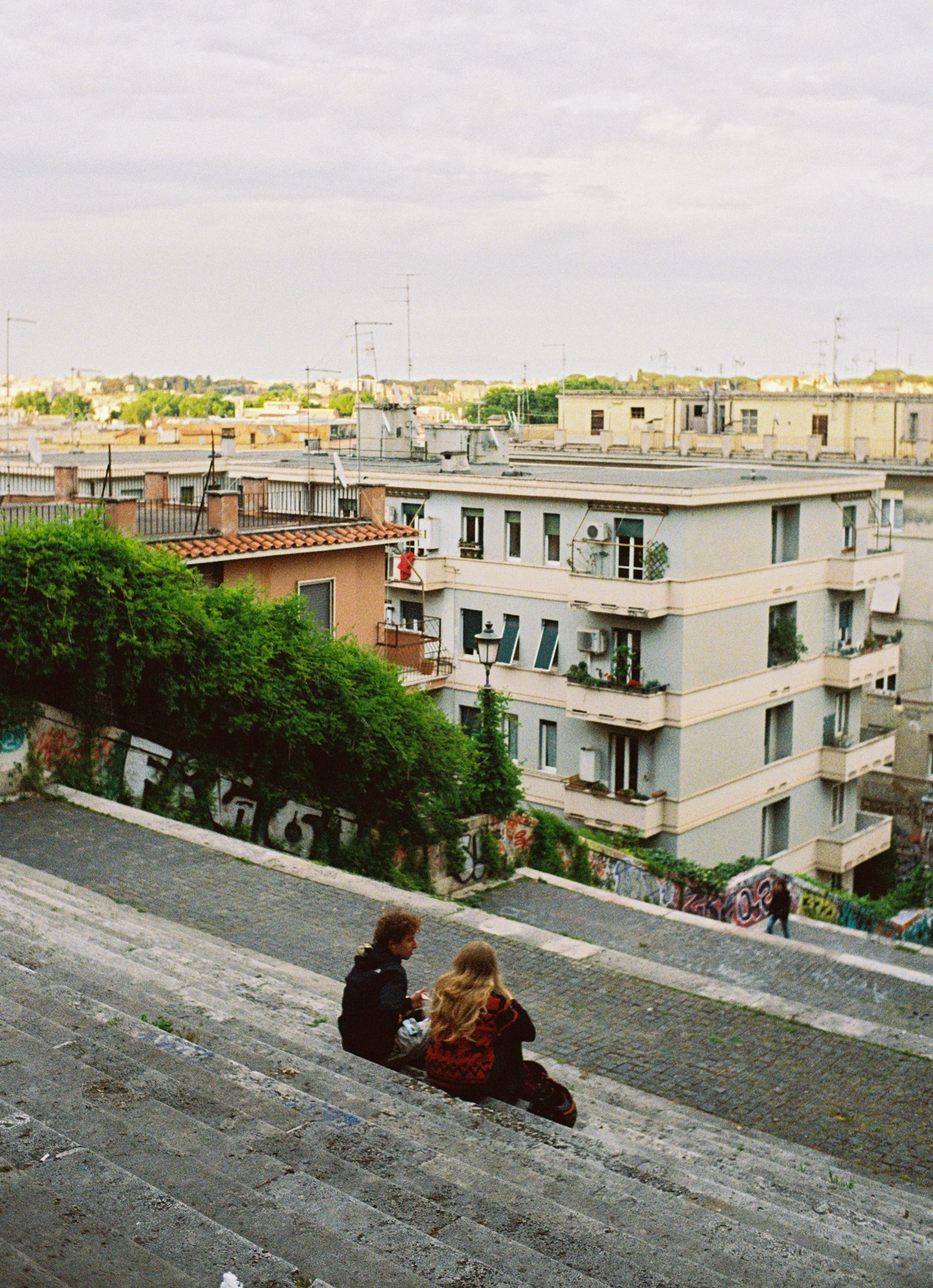 Two young people sit on a stone stairway in an urban area, overlooking a cityscape with apartment buildings, trees, and graffiti-covered walls. The sky is overcast.