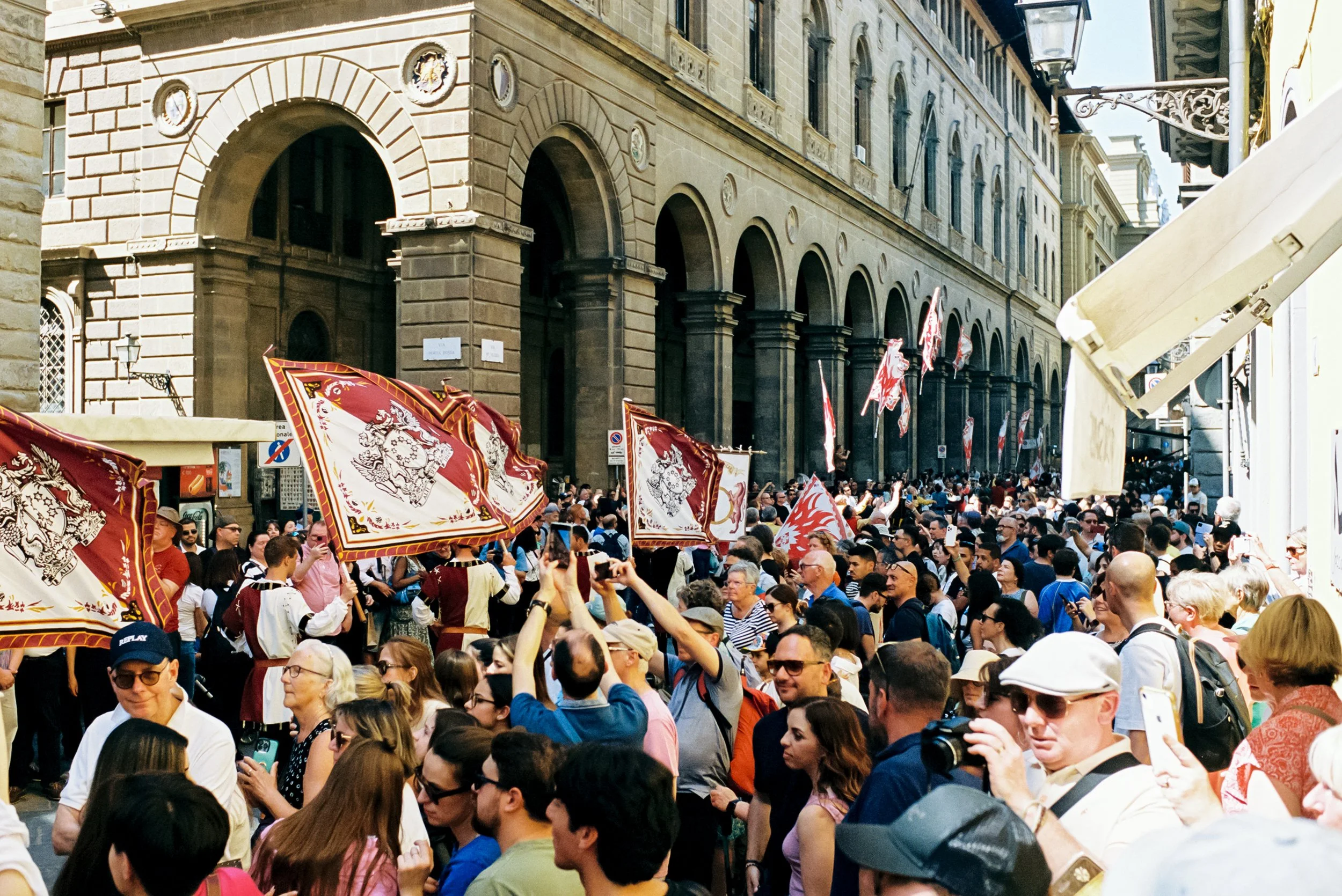 A large crowd of people gathered on a city street during a parade or festival, with people holding up flags, some taking photos, and historic buildings in the background.