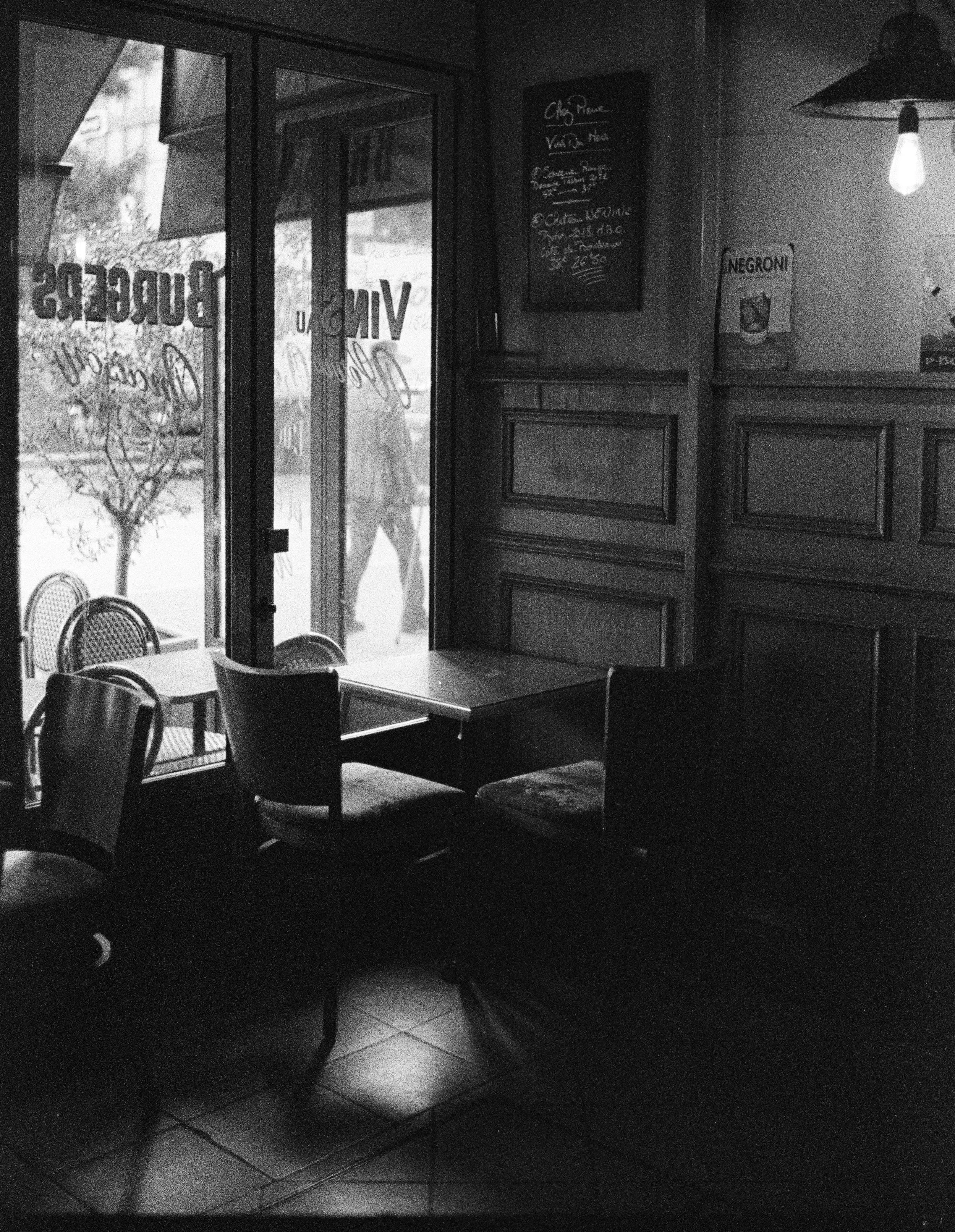 Interior of a quiet cafe with wooden paneling, a small table with chairs, and a door leading outside, showing sunlight and a person walking by.
