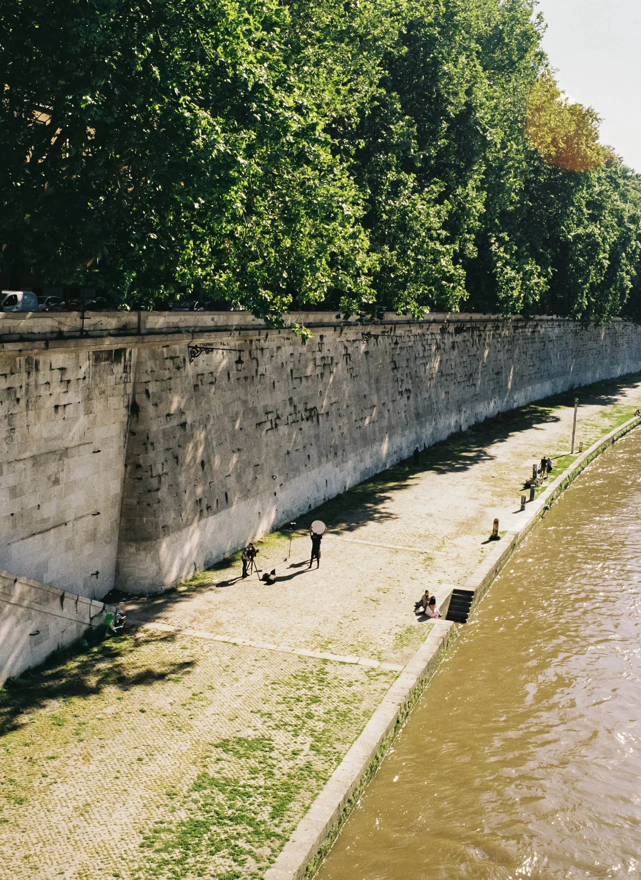 People sitting and walking along the riverbank with a stone wall and green trees overhead