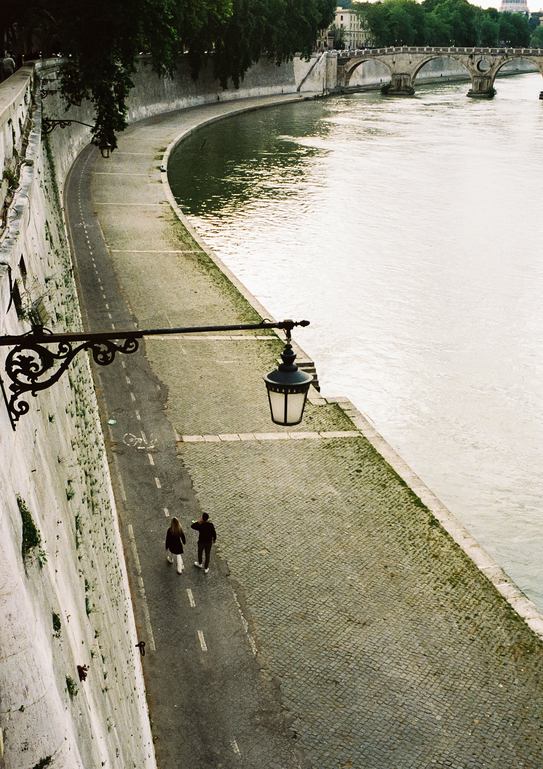 Two people walking along a riverside pathway with cobblestone pavement, lined with trees and old-style street lamps, next to a stone wall and a river with a bridge in the background.