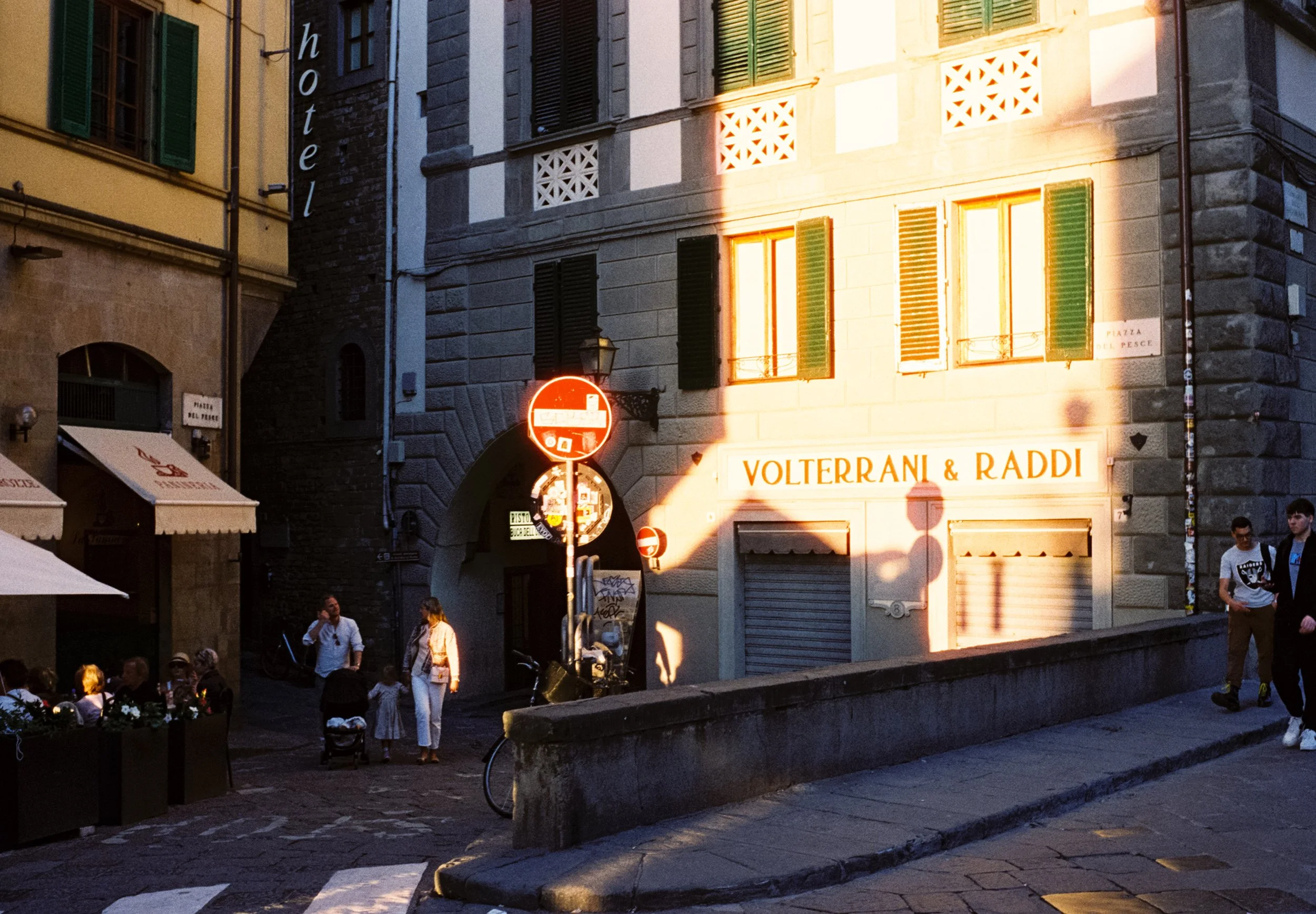 A street scene at sunset in an Italian city with buildings, pedestrians, and outdoor dining. The sunlight creates a warm glow on the buildings.