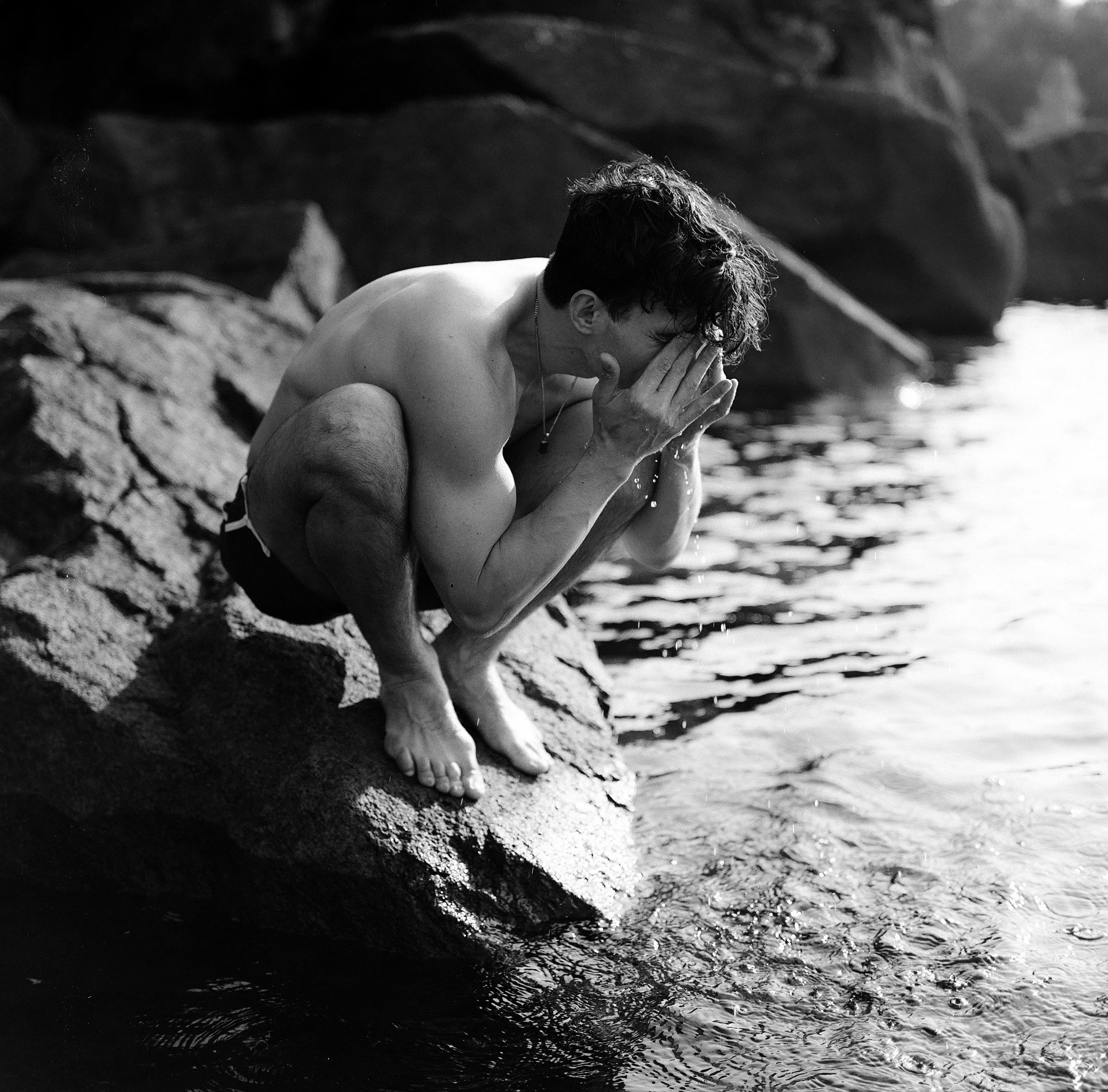 A black and white photo of a man squatting on a rock at the edge of water, washing his face with his hands.