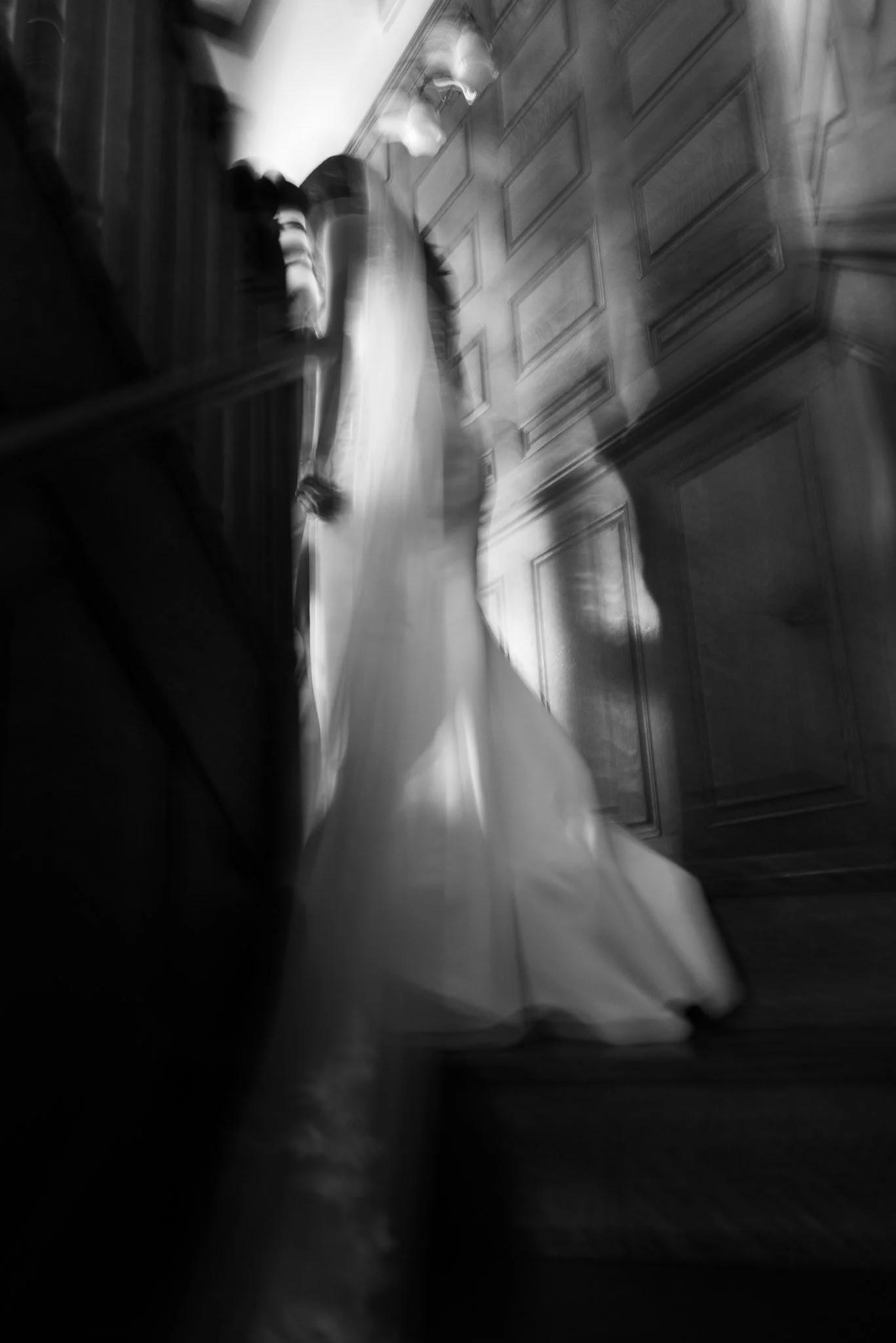 Blurred black and white photo of a person standing in front of a paneled wooden wall, wearing striped socks and a long skirt or dress.