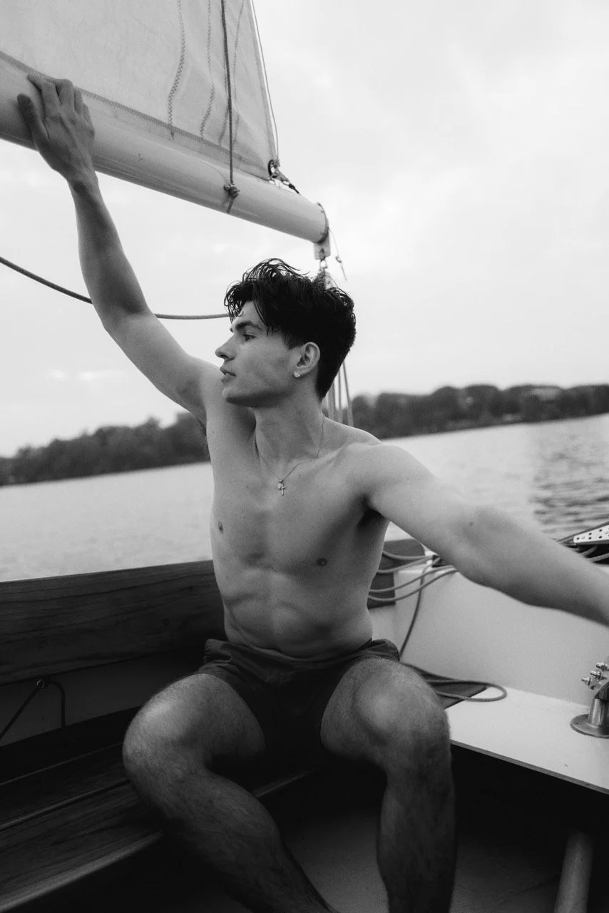 A young man with dark hair and a cross necklace, shirtless, sitting on a boat near a body of water, holding a sail, with a distant shoreline in the background, in black and white.