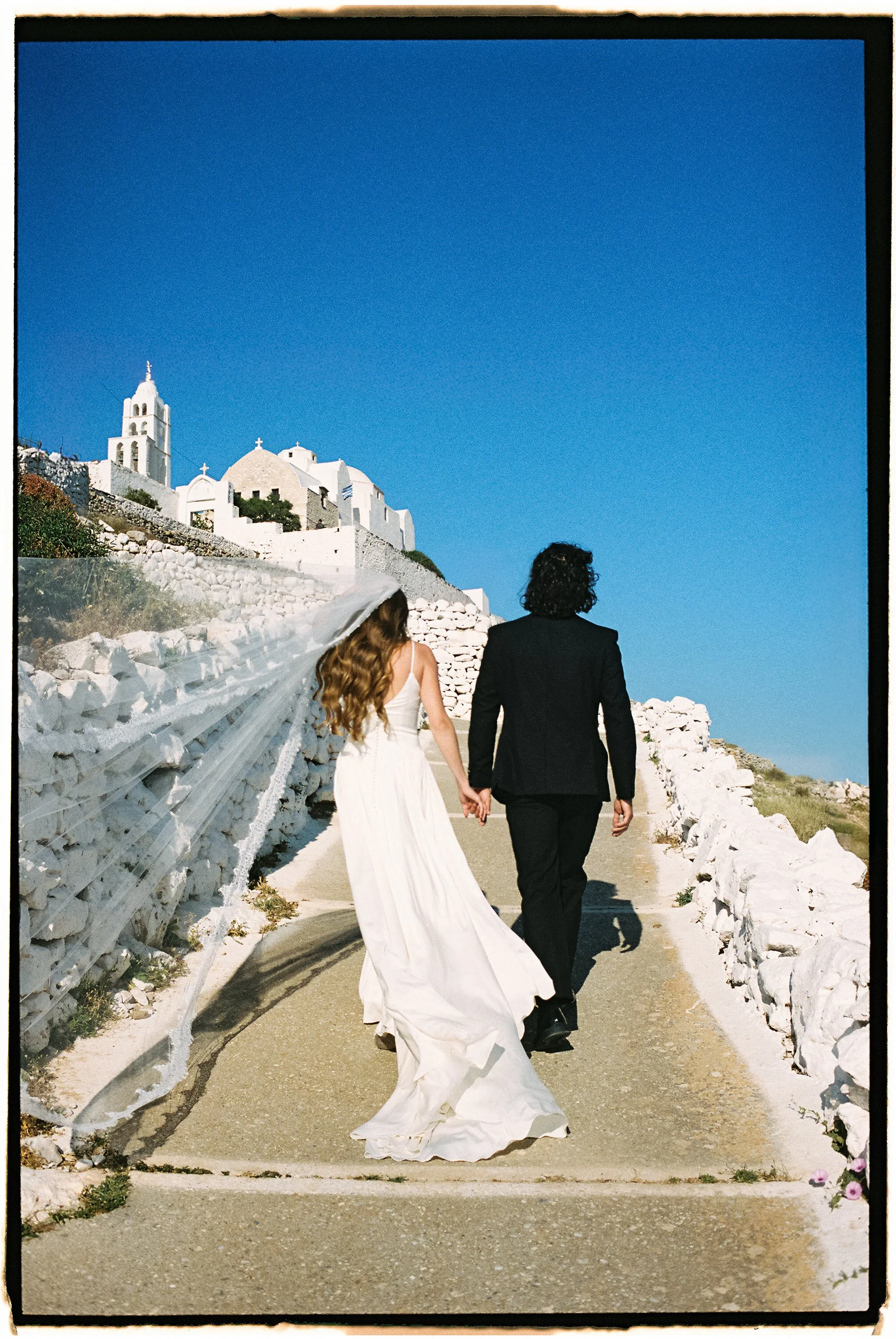 A bride and groom holding hands walking along a path in a coastal town with white buildings and blue sky in the background.